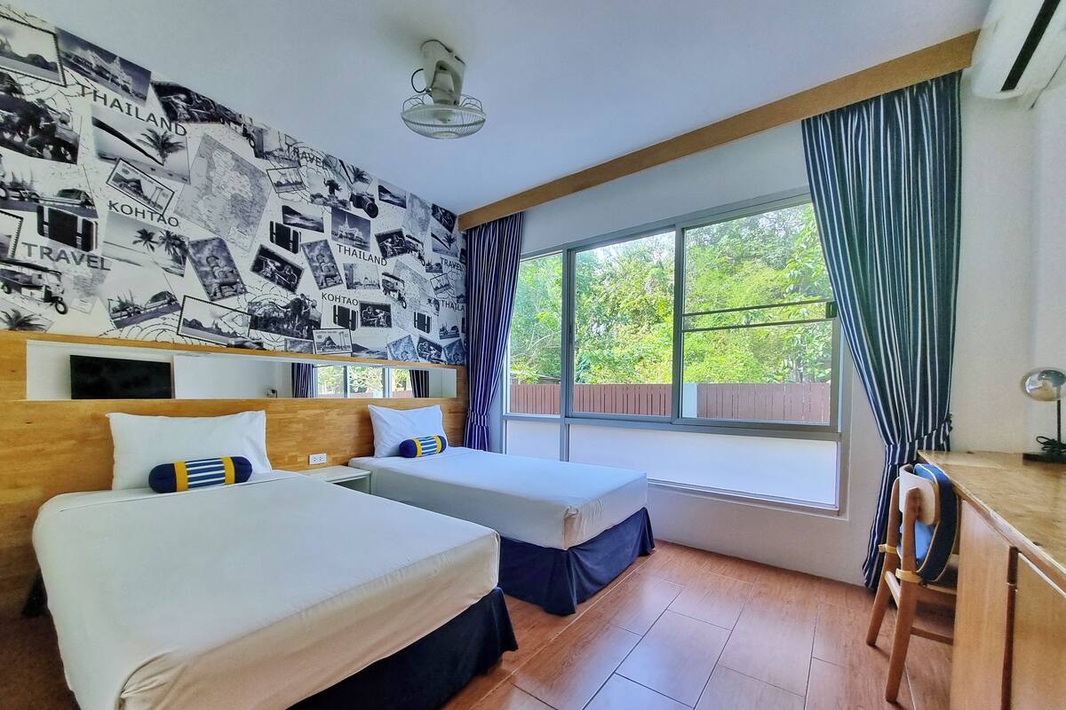 Une chambre d’hôtel avec deux lits, une tête de lit en bois, une grande fenêtre avec des rideaux et un mur décoré de photos de voyage.