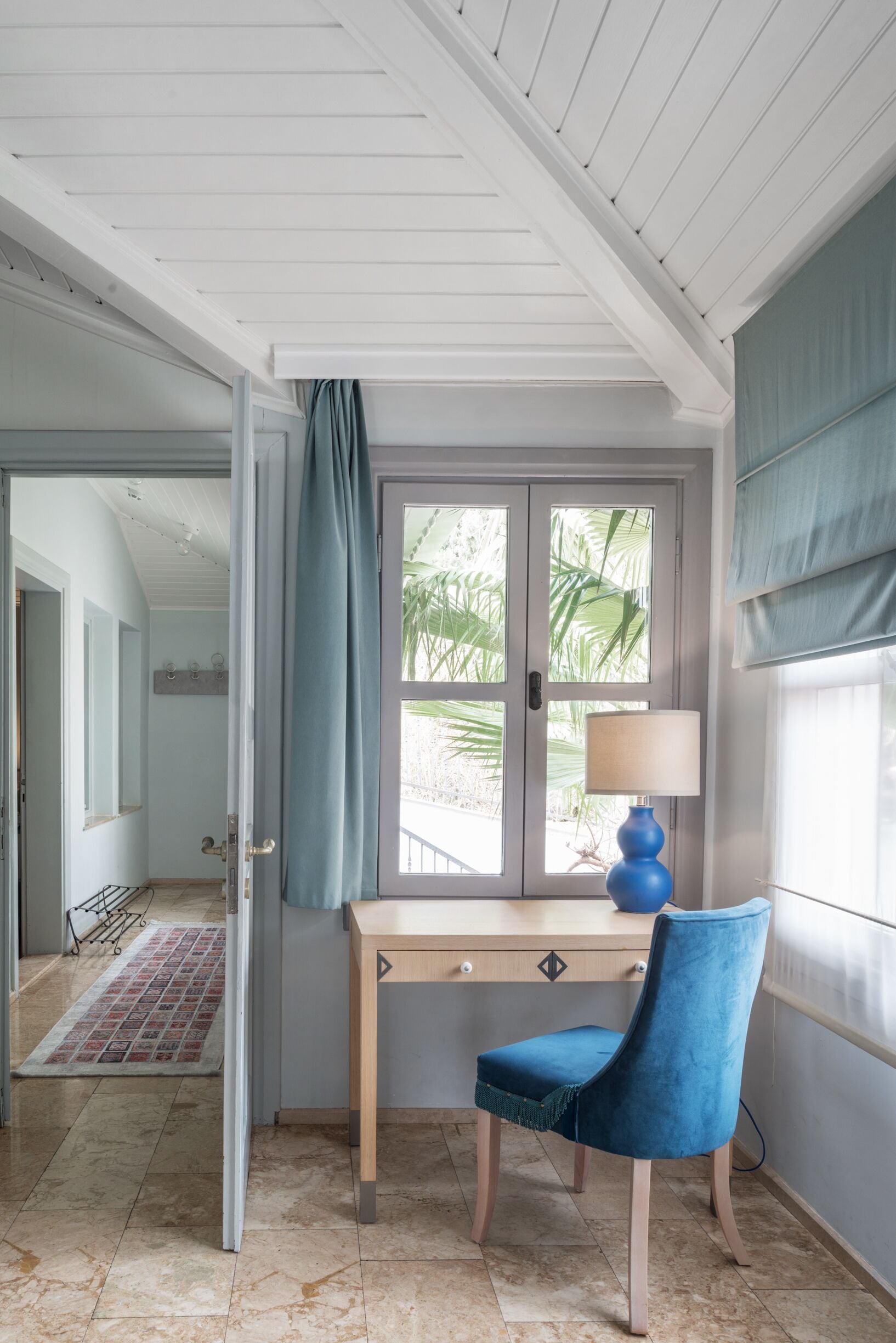Una habitación con un escritorio de madera, una silla azul, una lámpara y un ventanal con vistas a la vegetación.