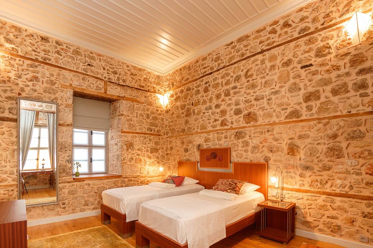 A room with stone walls, two beds, a wooden desk, a mirror, and a window with curtains.