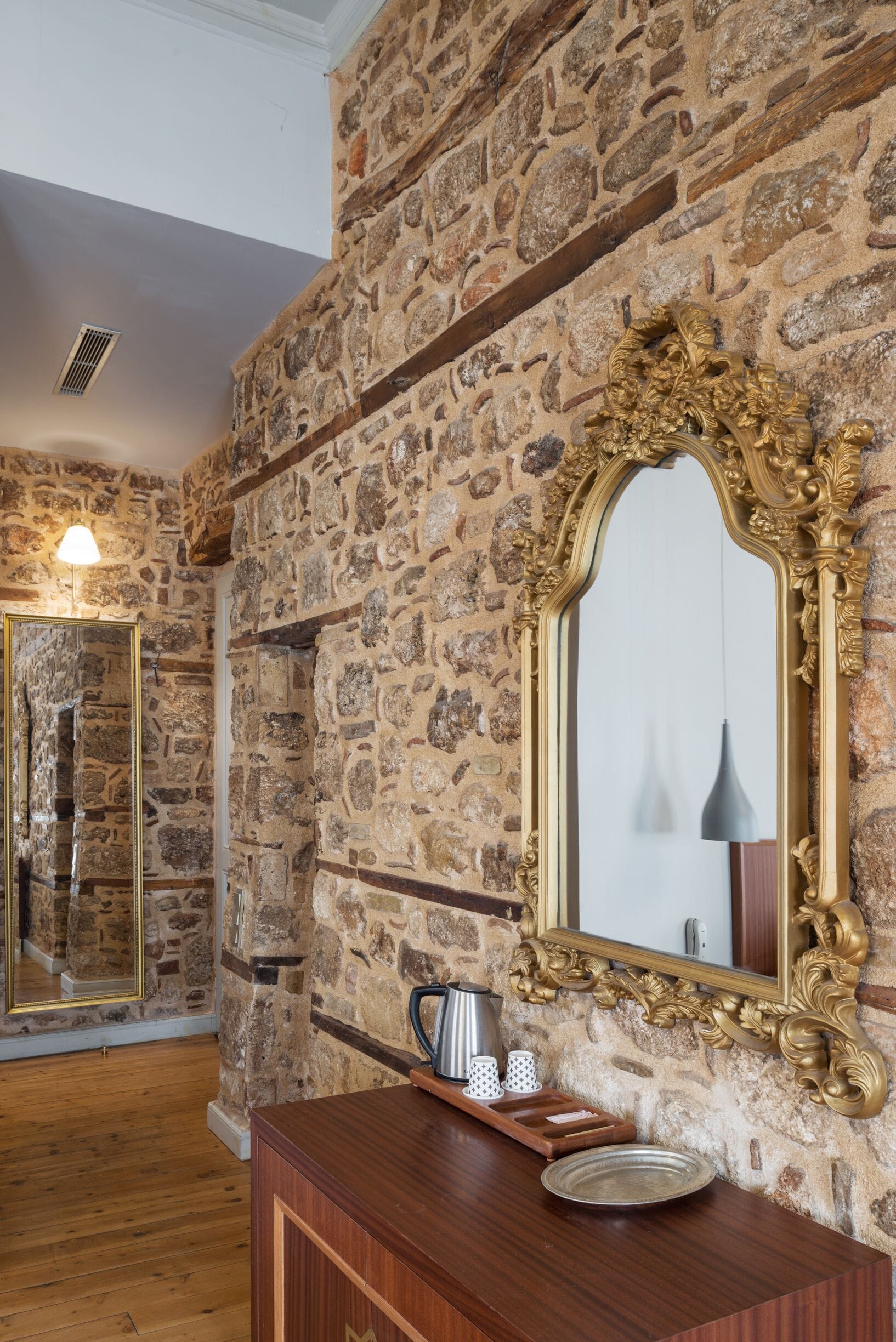A room with stone walls, a wooden floor, a mirror with an ornate frame, and a wooden cabinet with a tray.