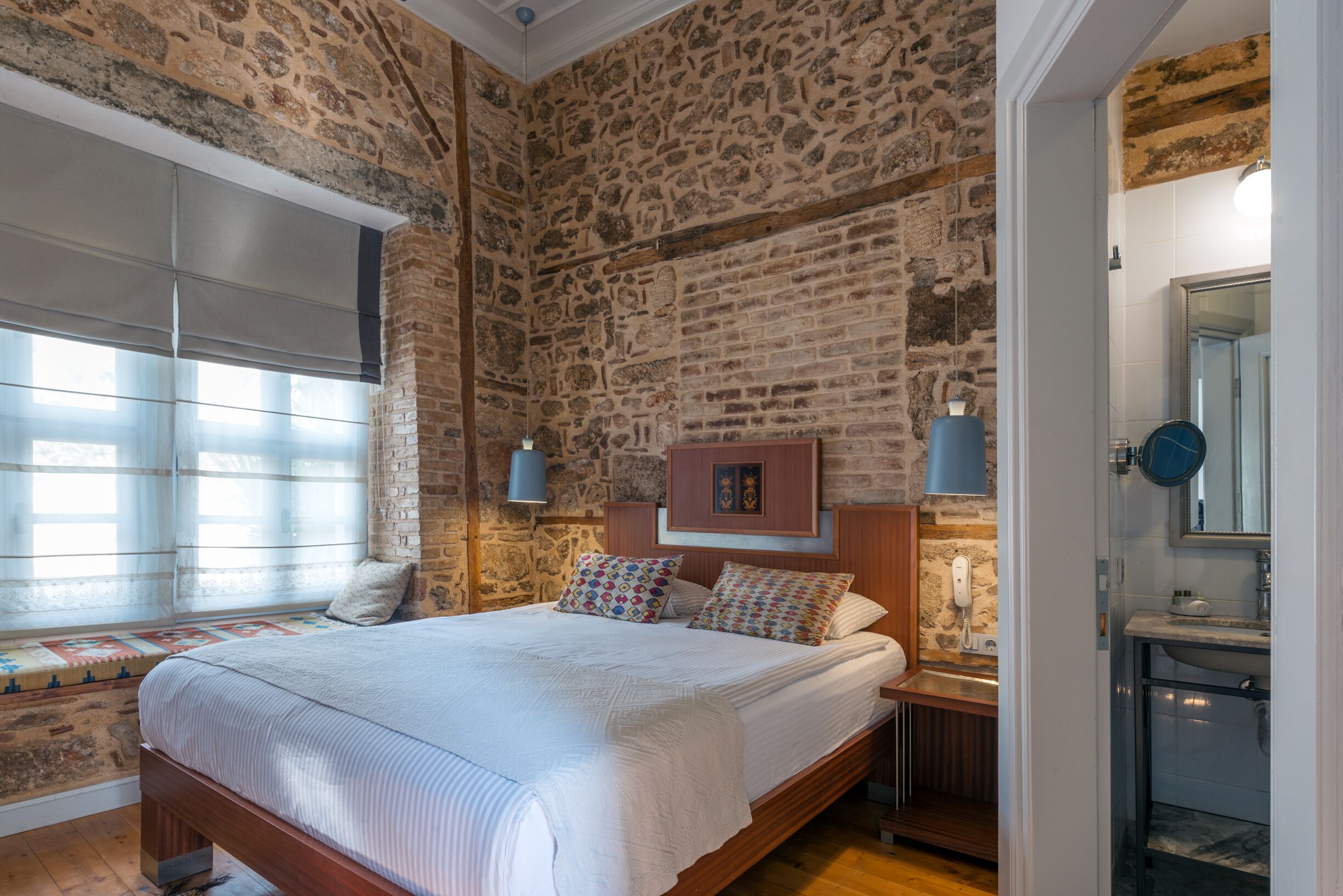 A bedroom with a stone wall, a large bed with patterned pillows, a bedside table with a lamp, and a bathroom with a mirror and sink.