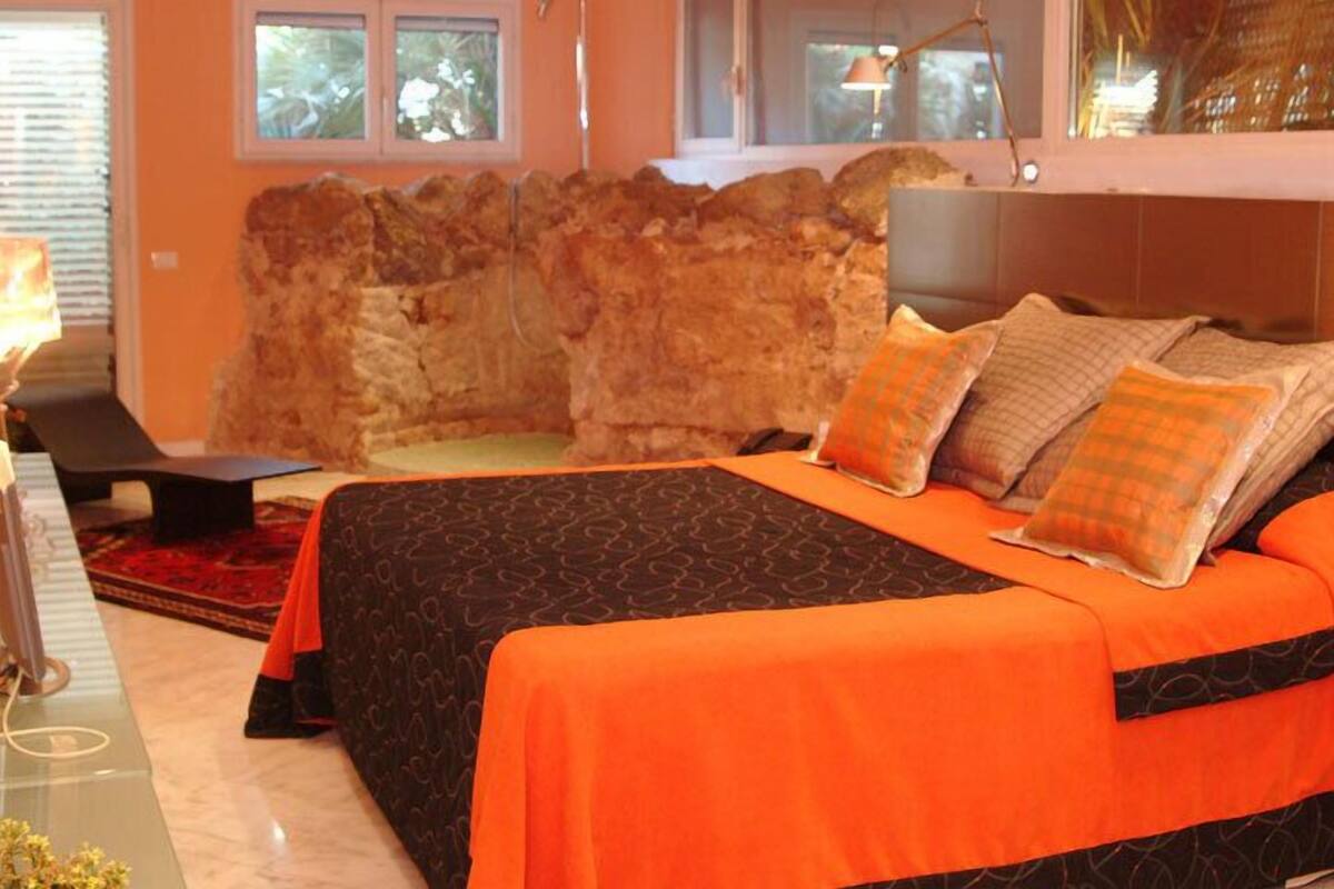 A hotel room with a large bed, a stone wall, a desk with a lamp, and a view of the outdoors.
