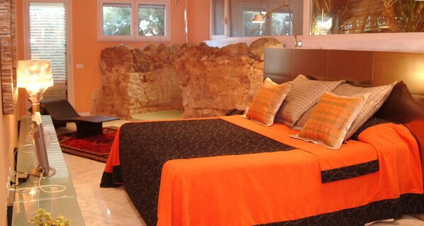 A hotel room with a large bed, a stone wall, a desk with a lamp, and a view of the outdoors.