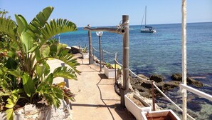 A coastal walkway with potted plants, a metal railing, and a clear view of the sea with a sailboat in the distance. - Le Stanze sul Mare (Carini)