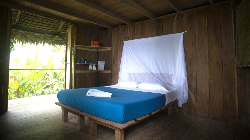 A wooden room with a bed, a shelf with toiletries, and a view of greenery outside.