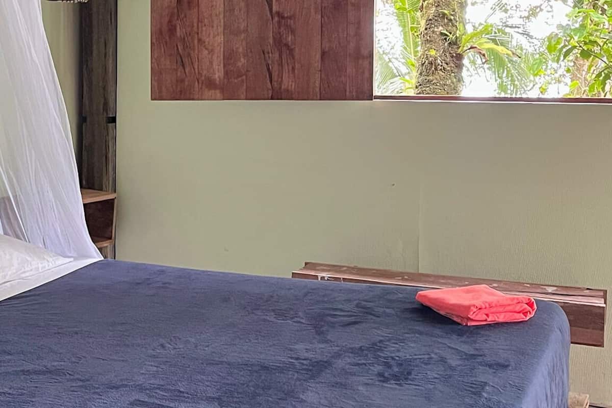 A bed with a blue bedspread and white pillows, a wooden headboard, and a window with a view of greenery.