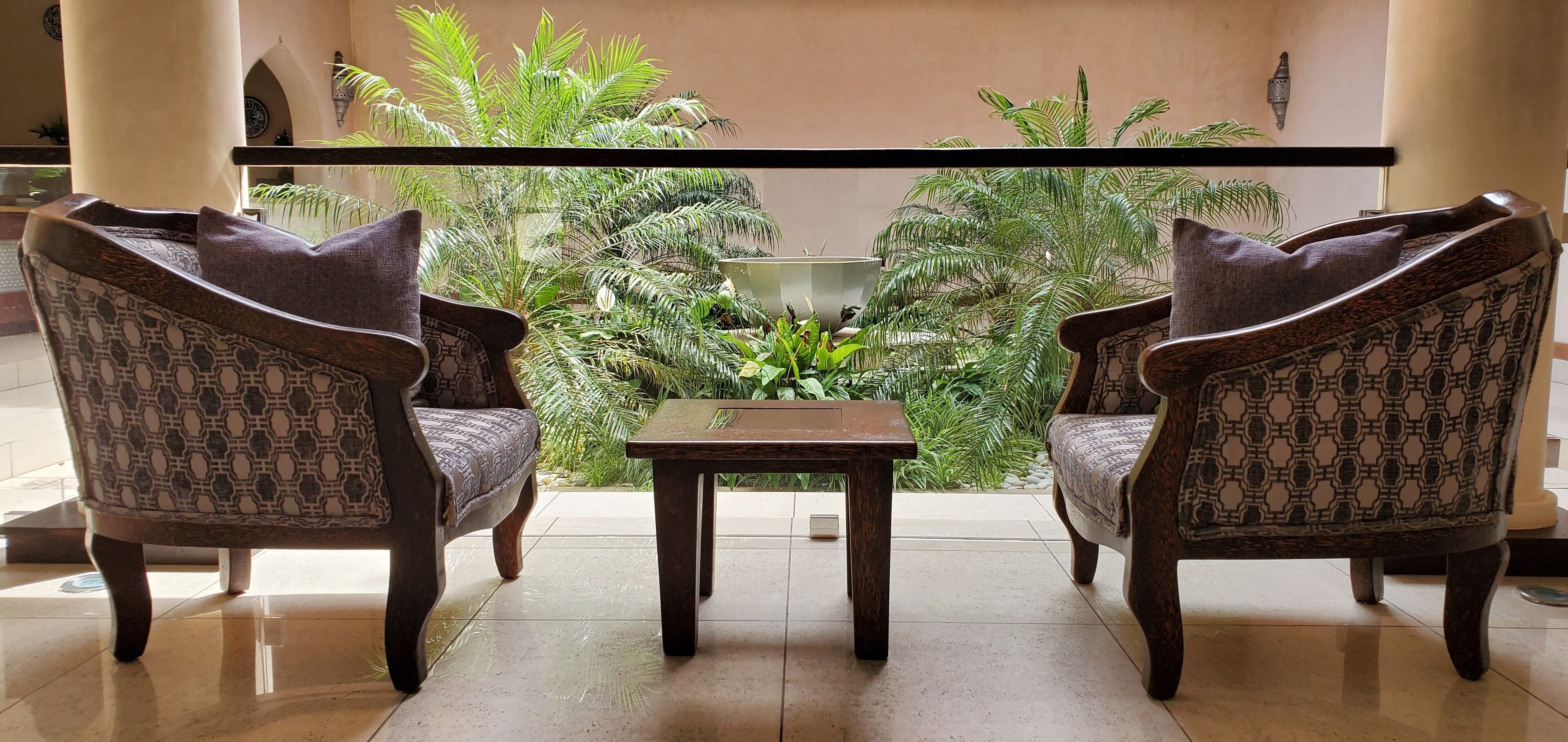 A seating area with two patterned chairs and a small table, surrounded by lush green plants and a decorative wall.