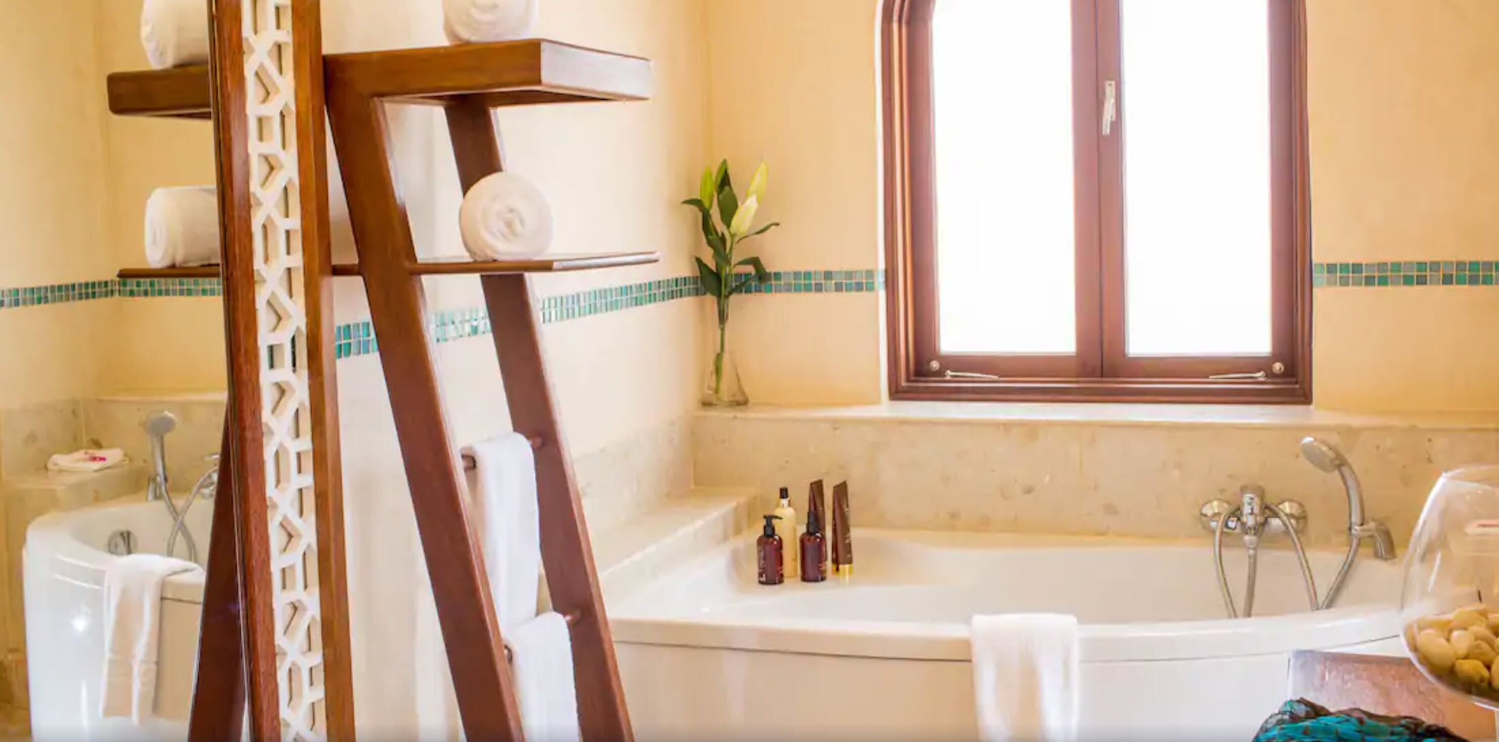 Un baño con bañera, ducha y ventana con vistas. Hay toallas en un perchero y artículos de aseo en el lavabo.