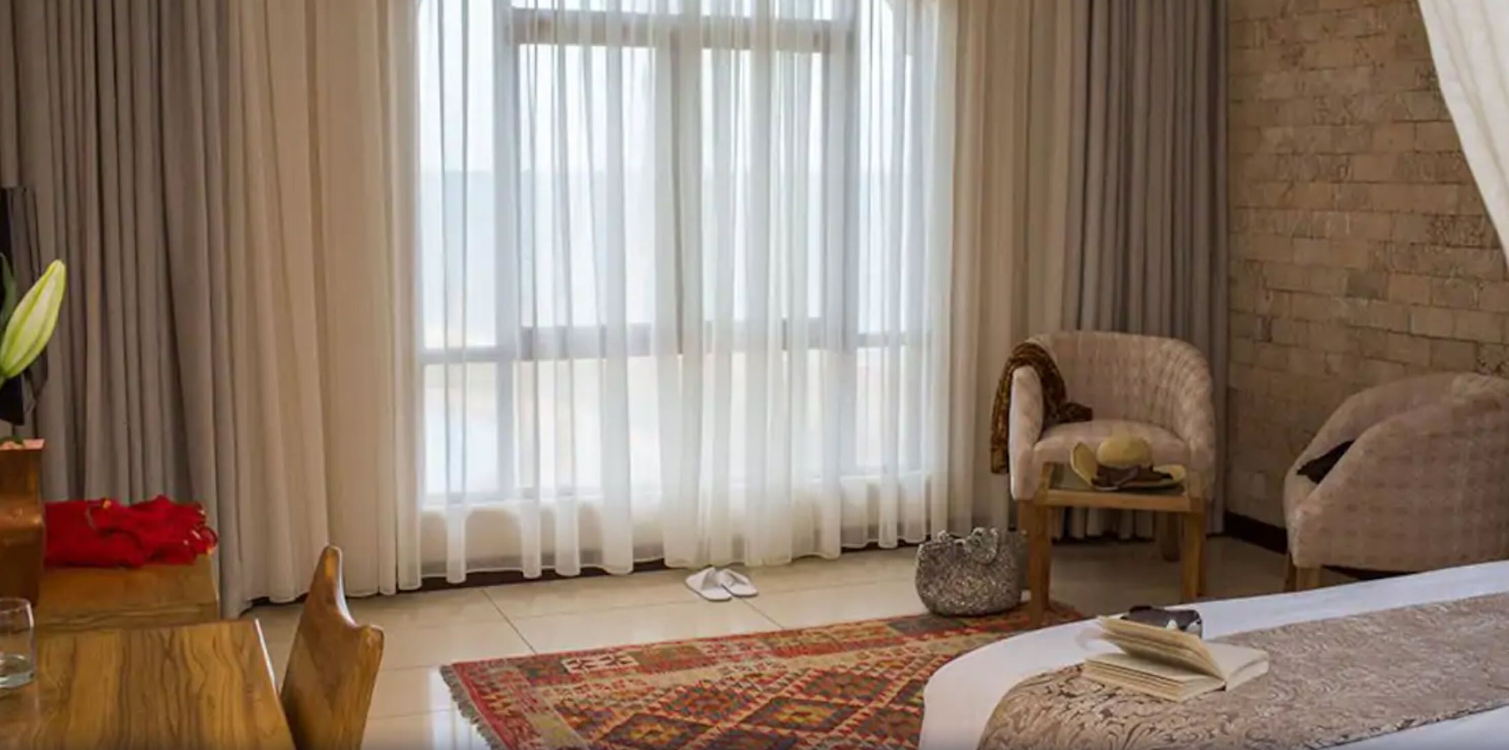 A hotel room with a large window, a bed, a chair, a small table, and a rug.