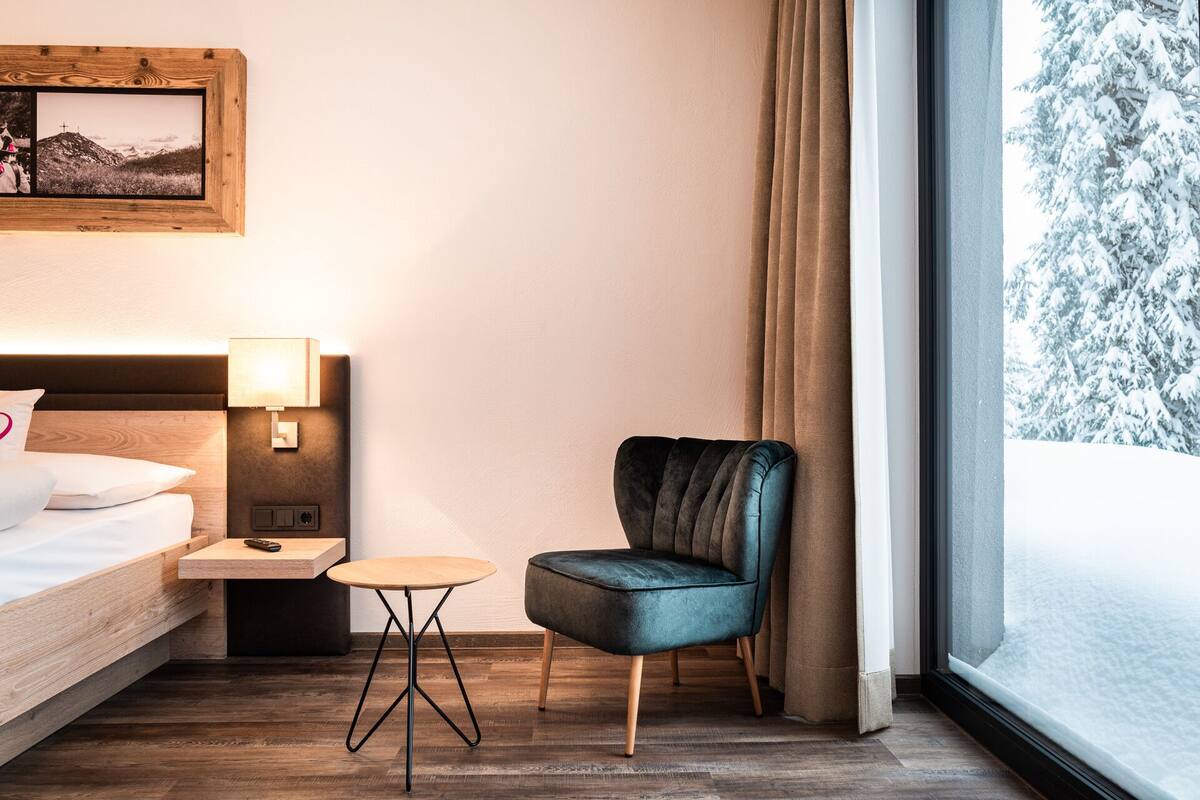 Habitación de hotel con cama, una mesita, una silla, una mesita de noche, una lámpara, un marco de fotos y una ventana con vista nevada.