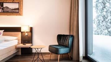 A hotel room with a bed, a small table, a chair, a nightstand, a lamp, a picture frame, and a window with a snowy view.