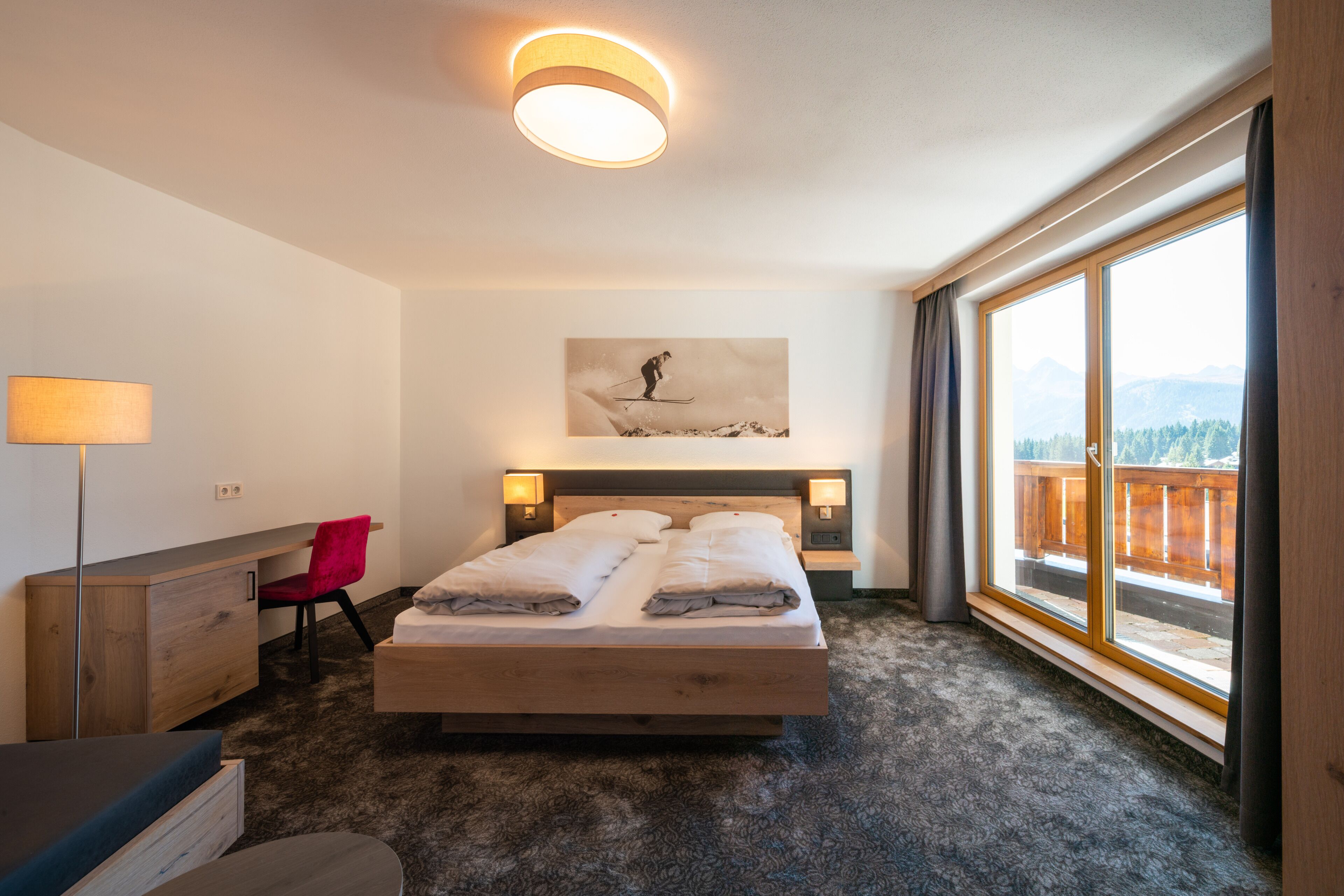 A bedroom with a bed, desk, chair, and large windows.