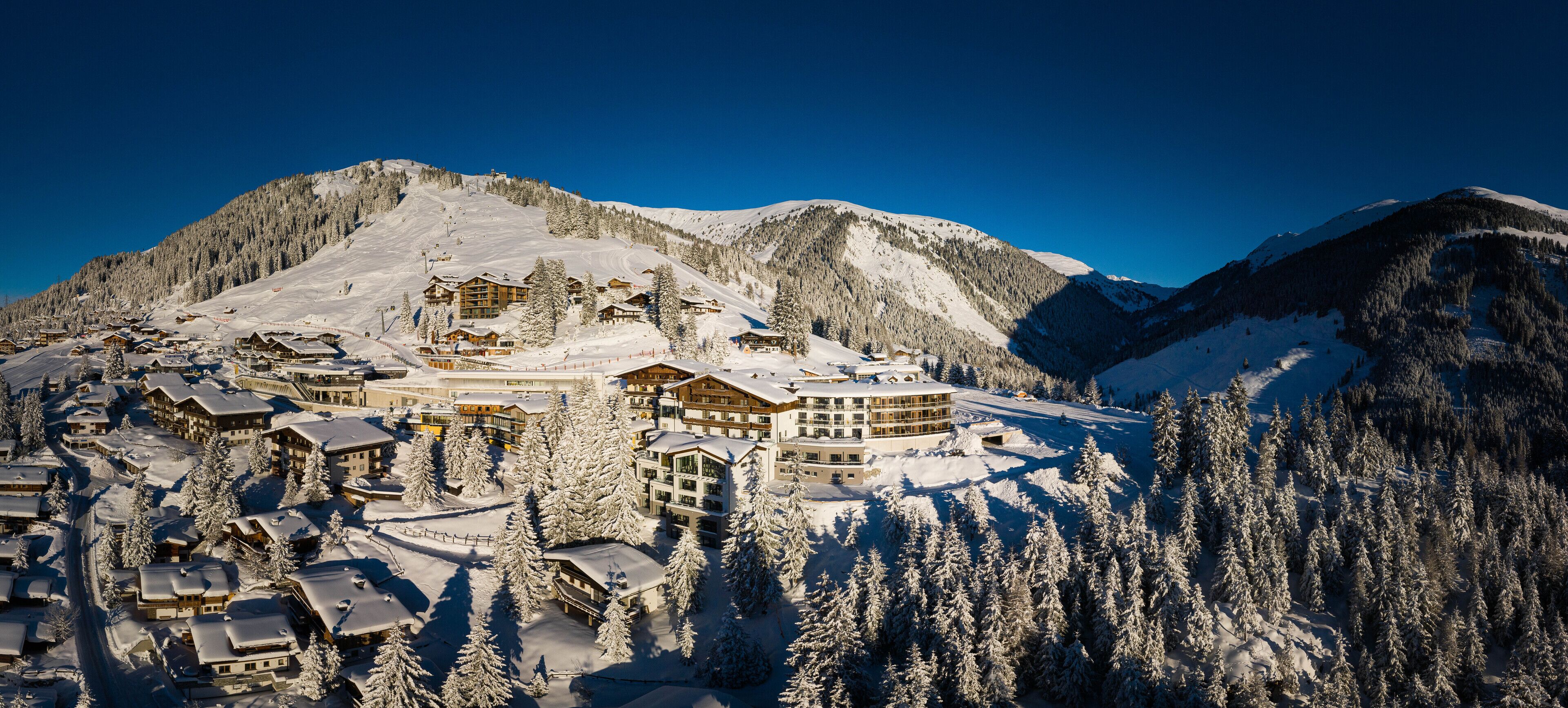 A snow-covered mountain resort with multiple chalet-style buildings, ski slopes, and dense forested areas.
