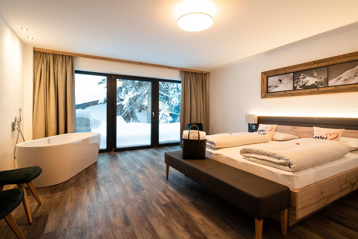 Un dormitorio moderno con una cama grande, un banco de madera, una bañera y vistas a un paisaje nevado a través de la puerta de cristal.