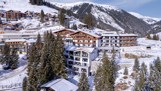Un complejo turístico en la montaña cubierto de nieve con edificios modernos y tradicionales, incluyendo estructuras de varios pisos y cabañas.