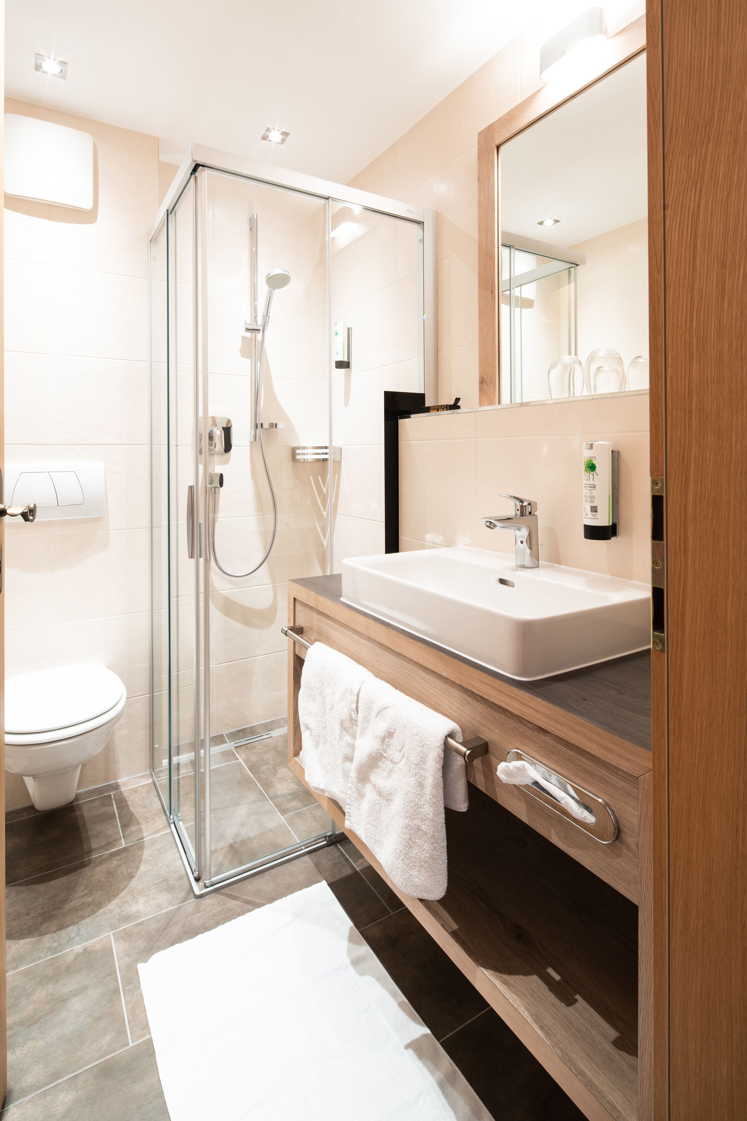 Un baño moderno con ducha envuelta en vidrio, un lavabo blanco y un mueble de tocador de madera.