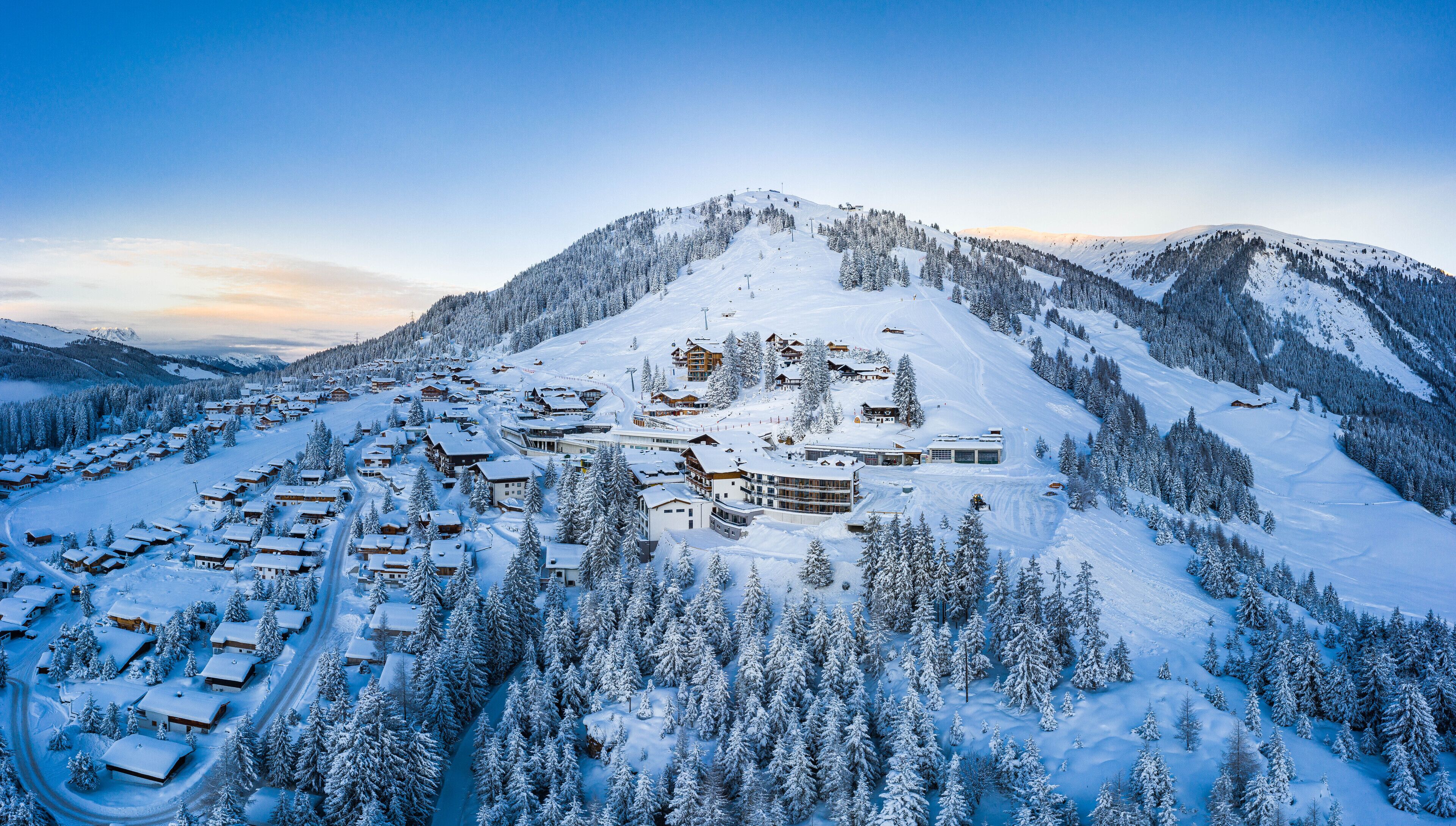A snow-covered mountain resort with multiple buildings, including lodges and a large hotel, surrounded by dense forests.