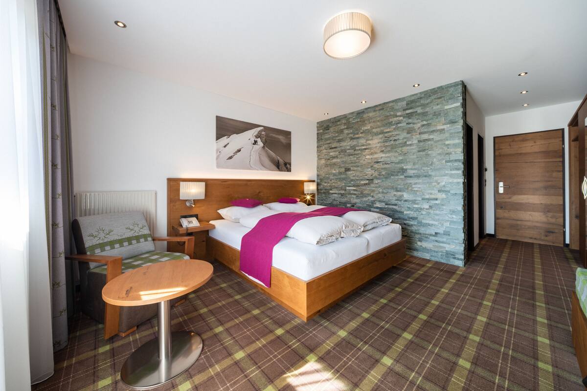 Habitación de hotel con cama, silla, mesita pequeña, mesita de noche, lámpara de pared, cuadro y pared de piedra.