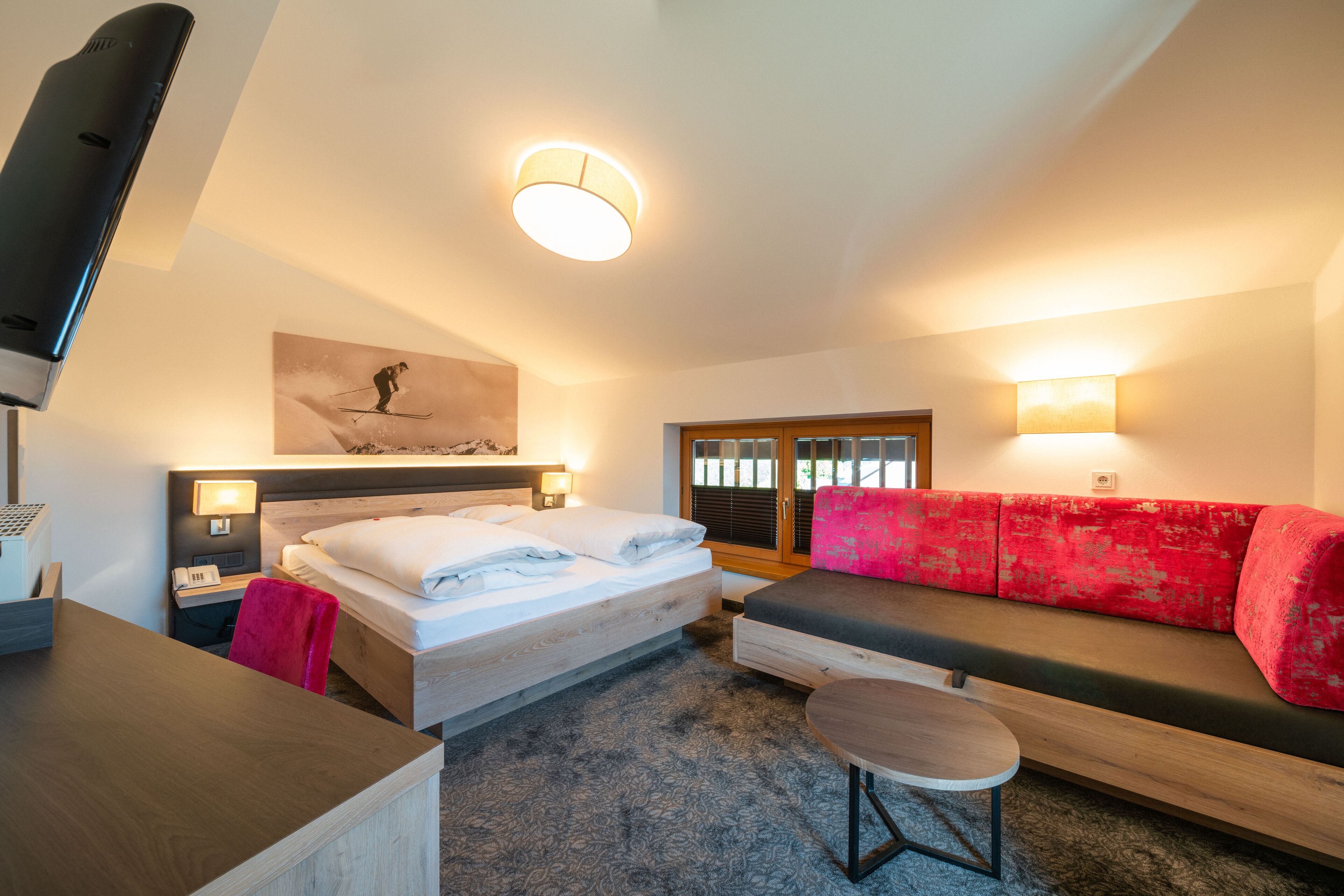 A modern hotel room with a bed, a red sofa, a small table, and a ceiling light.