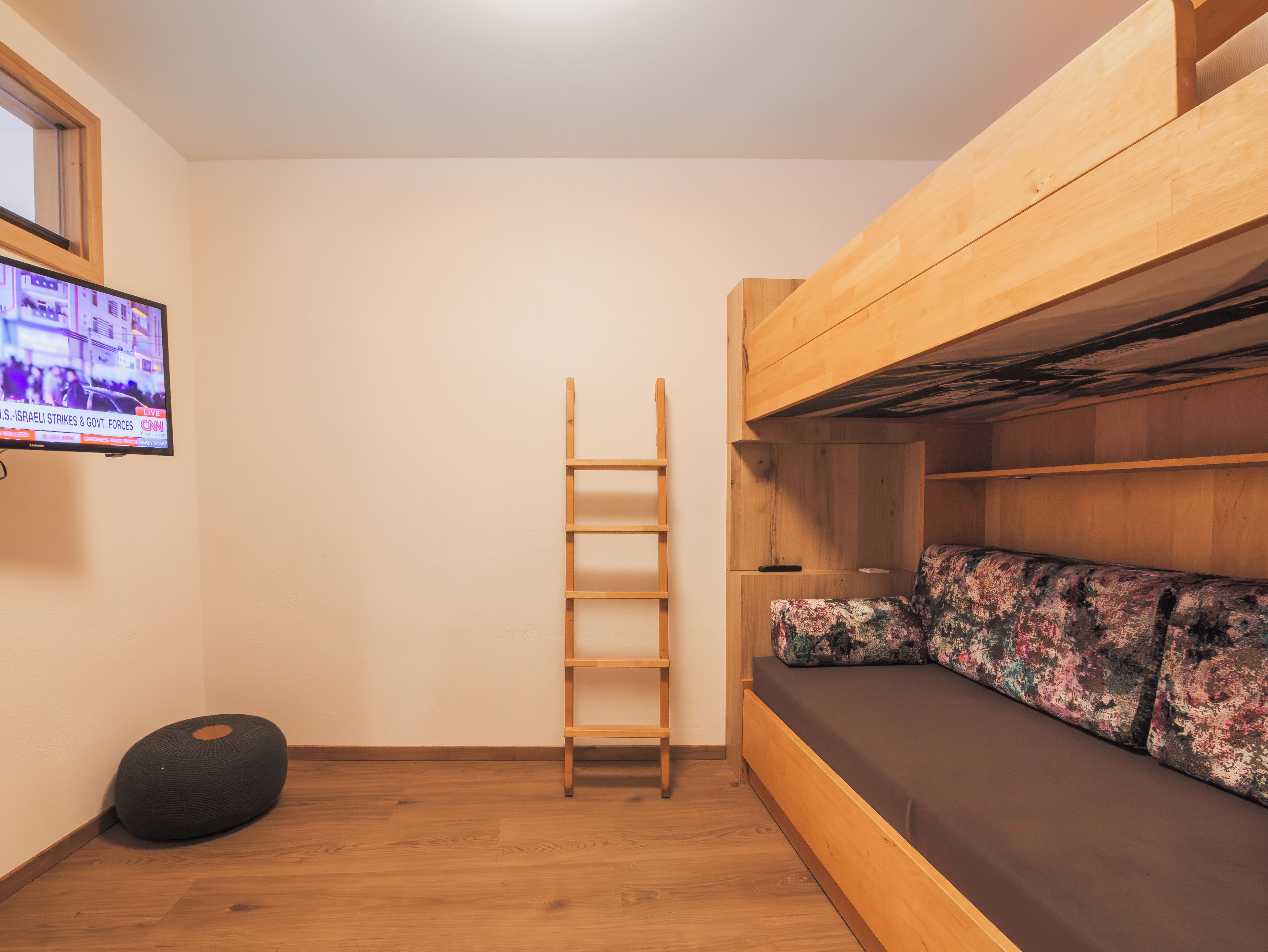 Een slaapkamer met stapelbed, een houten trapje, een televisie aan de muur en een zwart poef.