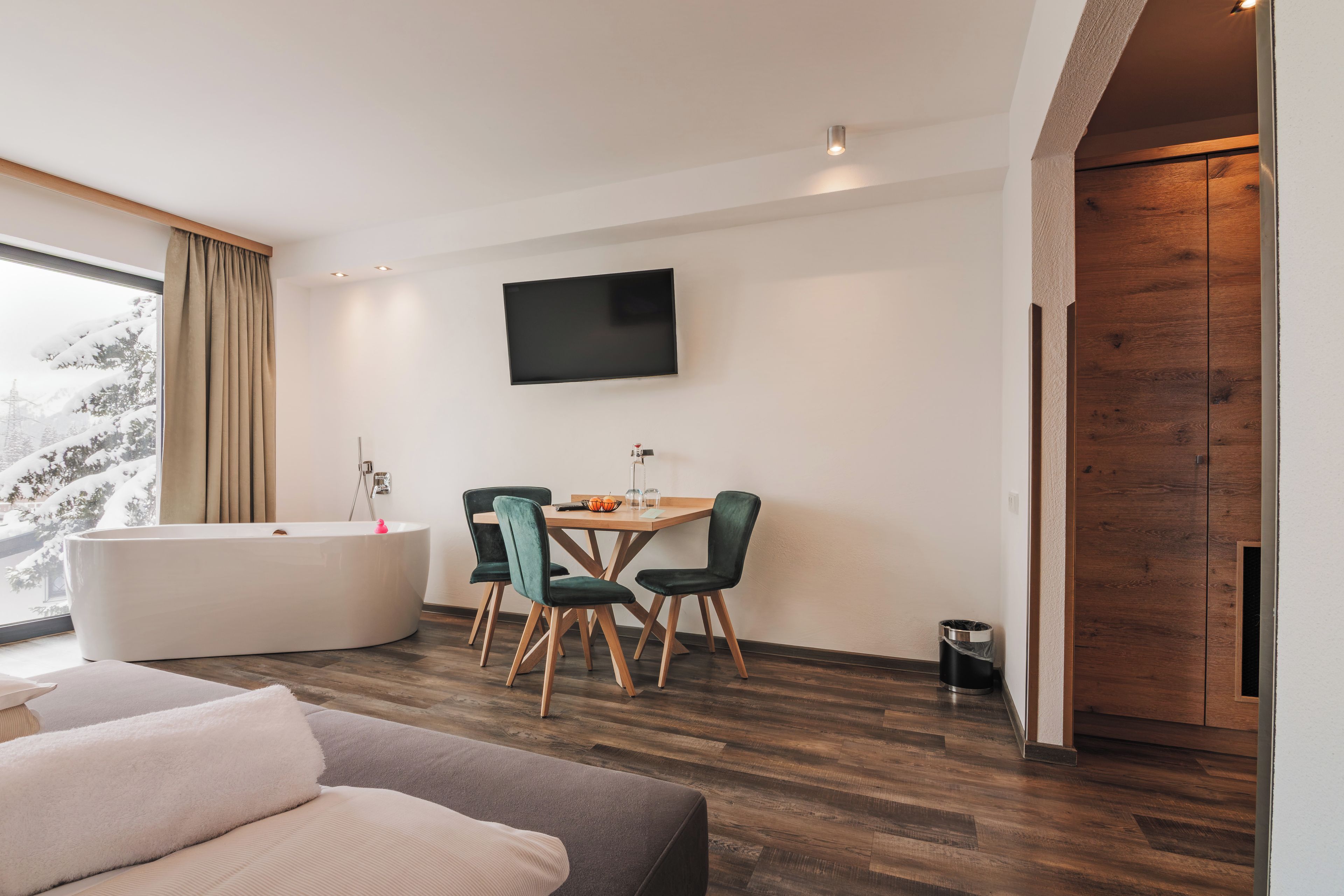Ein modernes Zimmer mit großem Fenster, einem Flachbildfernseher, einem Holzesstisch mit Stühlen, einer Badewanne und einem Sofa.