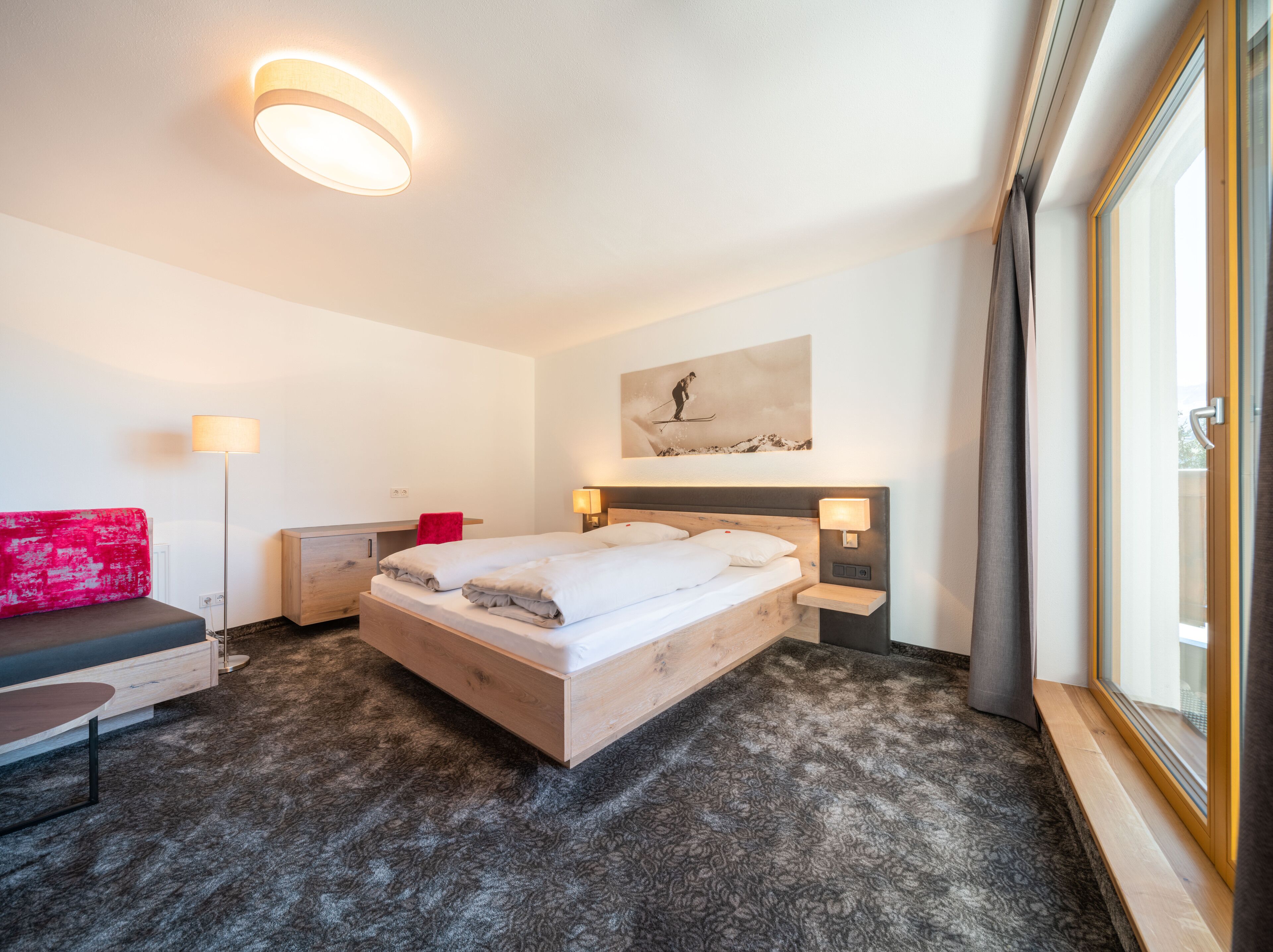 Une chambre d’hôtel avec un grand lit, un bureau, une chaise, un canapé rouge et une fenêtre avec des rideaux.