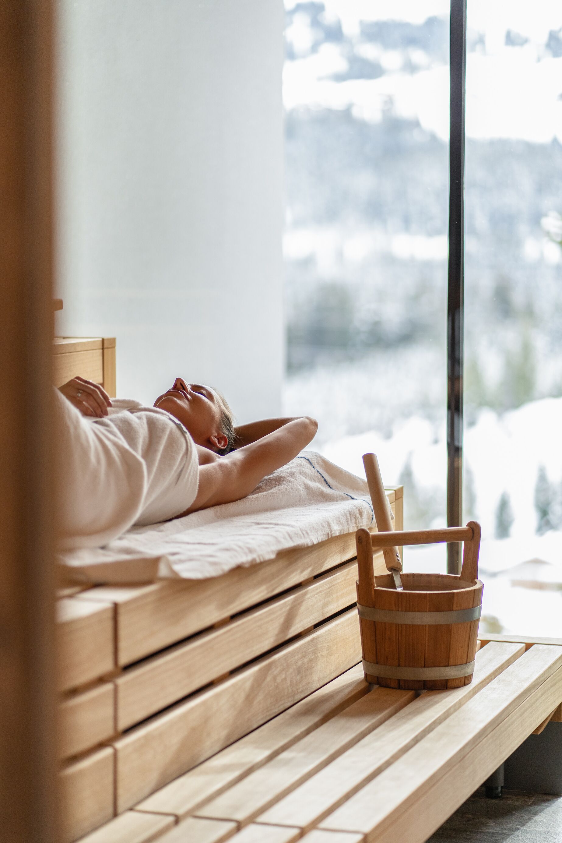 Eine Person liegt auf einer Holzbank, neben ihr ein Holzeimer. Im Raum befindet sich ein Fenster mit Blick auf eine verschneite Landschaft.