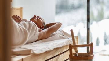 A person lying on a wooden bench with a wooden bucket beside them, in a room with a window overlooking a snowy landscape.