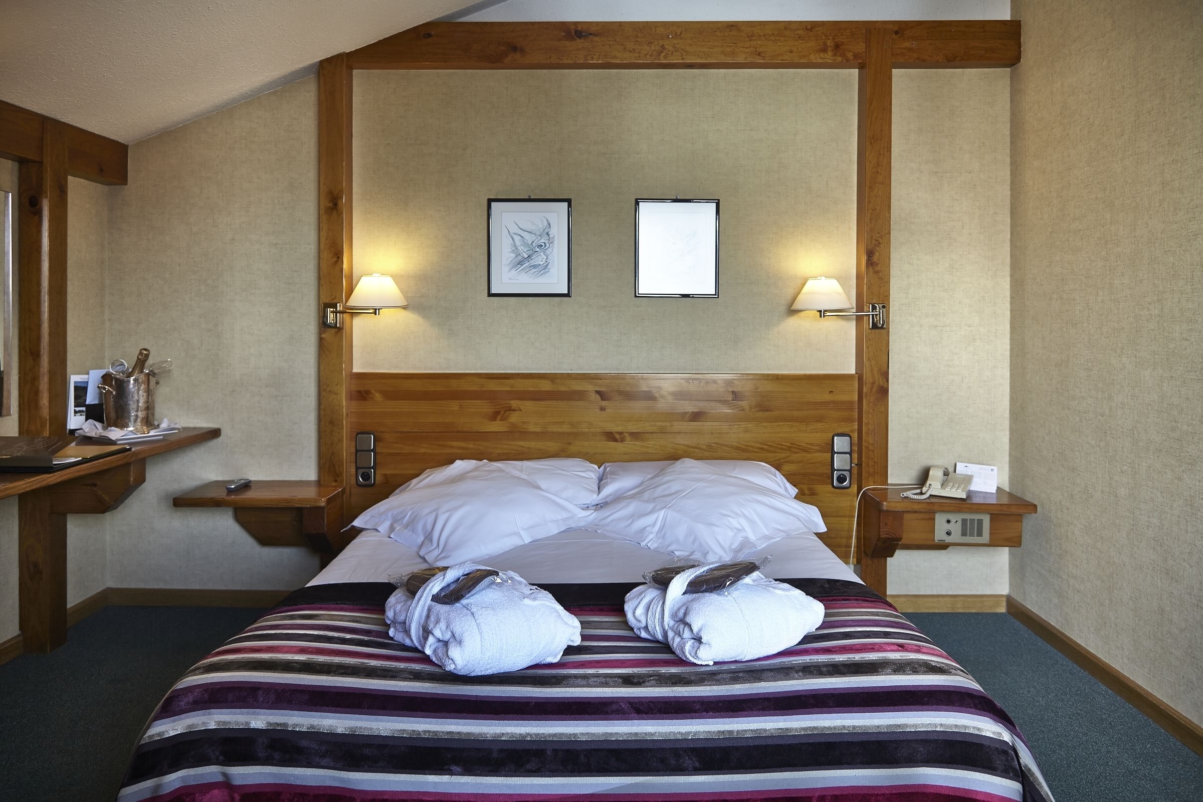 Hotelkamer met houten hoofdeinde, twee ingekaderde foto's, een nachtkastje met een lamp, een telefoon en een bed met gestreept beddengoed en handdoeken.