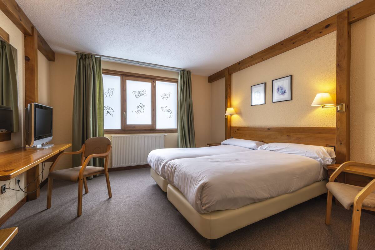 A hotel room with a wooden desk, a bed, two chairs, a television, and a window with curtains.