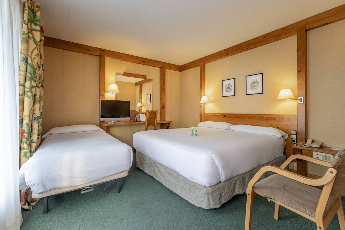 A hotel room with two beds, a chair, a desk with a lamp, and a television.