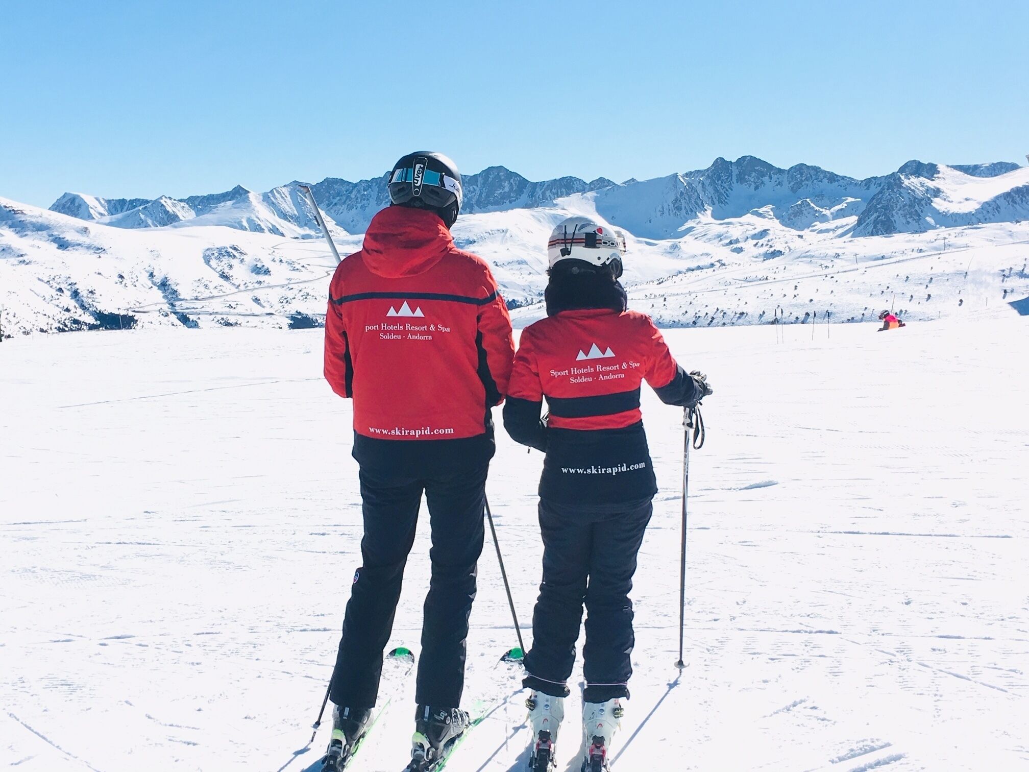 Two people skiing on a snowy slope with mountains in the background.