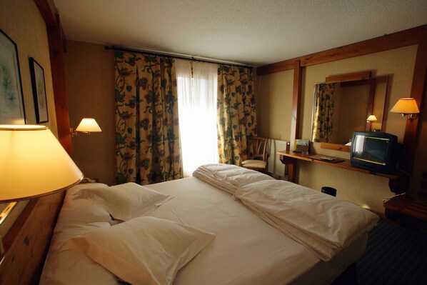A hotel room with a bed, a chair, a desk with a lamp, a television, and floral curtains.