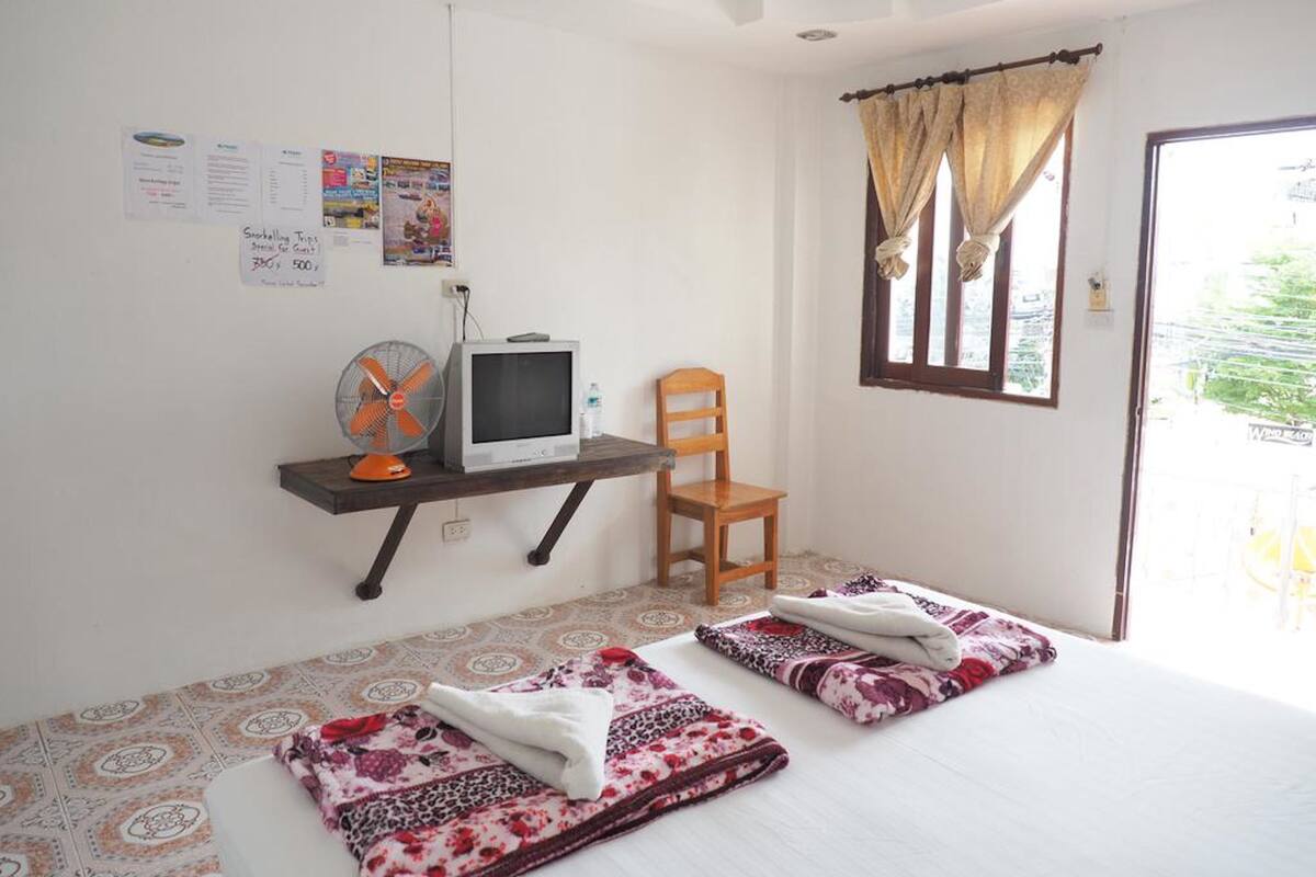 A small room with a bed, a chair, a desk with a computer and fan, and a window with curtains.