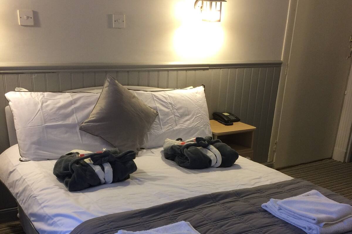 Quarto de hotel com cama, almofadas, mesa de cabeceira com telefone e luminária na parede.