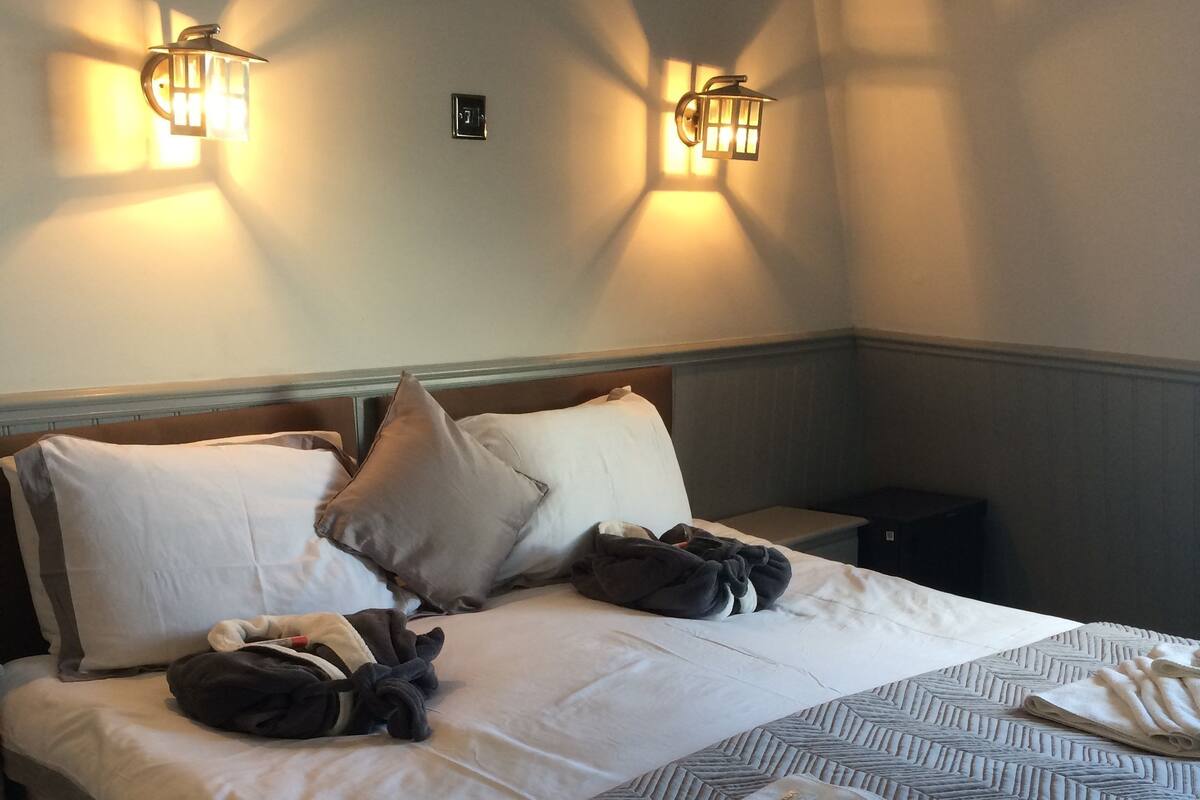 Cama bem arrumada com almofadas e toalha dobrada, duas luminárias fixadas na parede e uma mesinha de cabeceira.