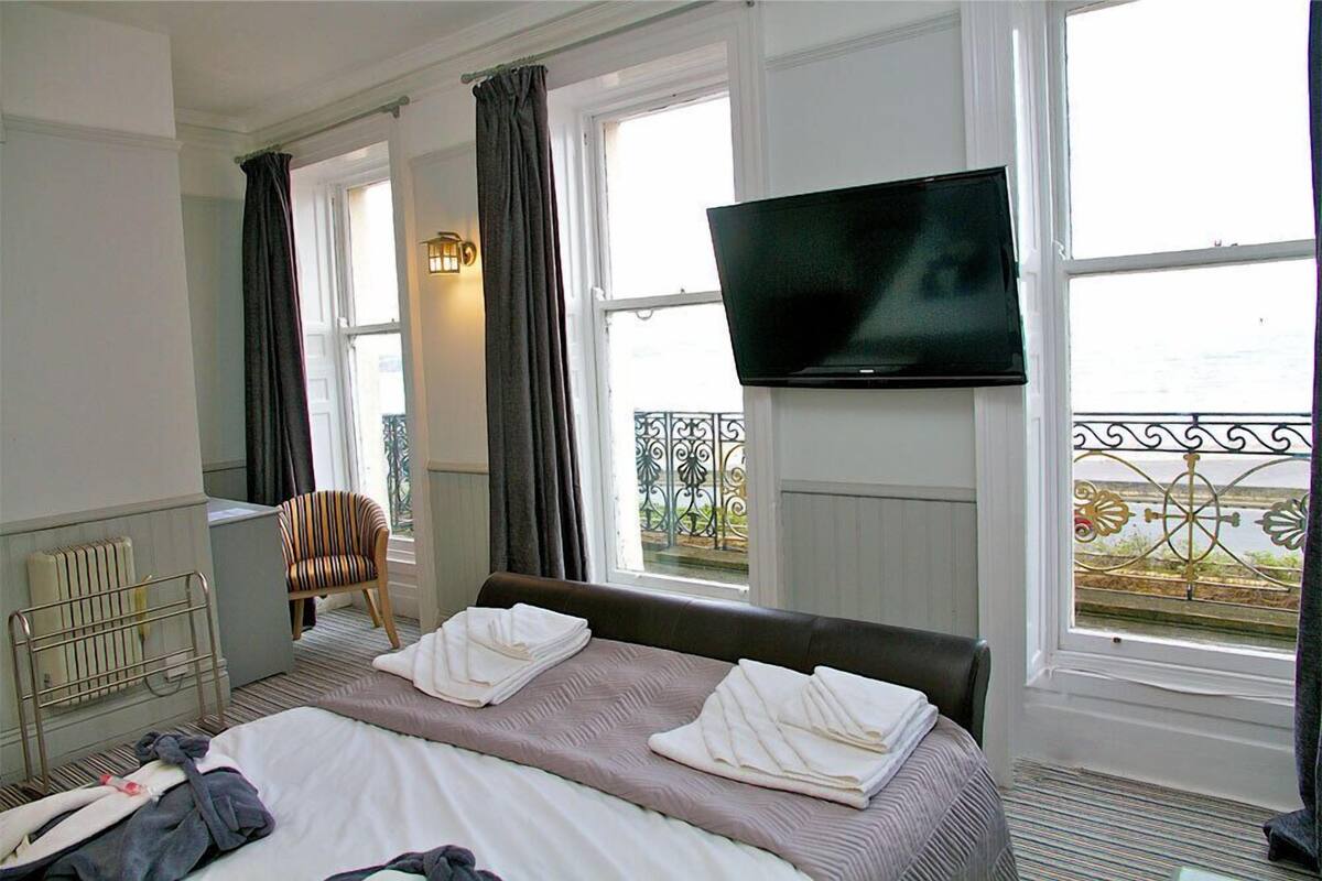 Quarto de hotel com cama, televisão na parede, uma cadeira e janela com vista.