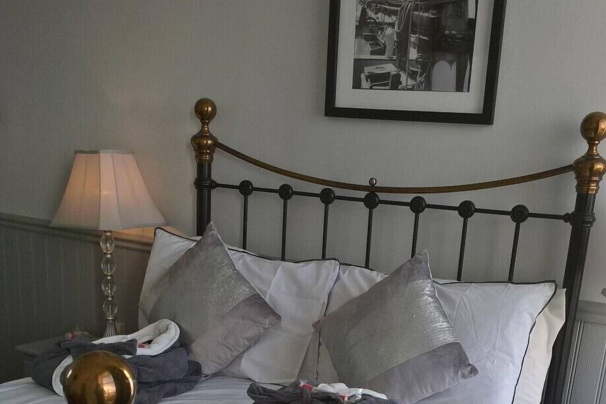 Cama bem arrumada com estrutura de metal, roupa de cama cinzenta e várias almofadas. Um quadro emoldurado está pendurado na parede acima da cama.