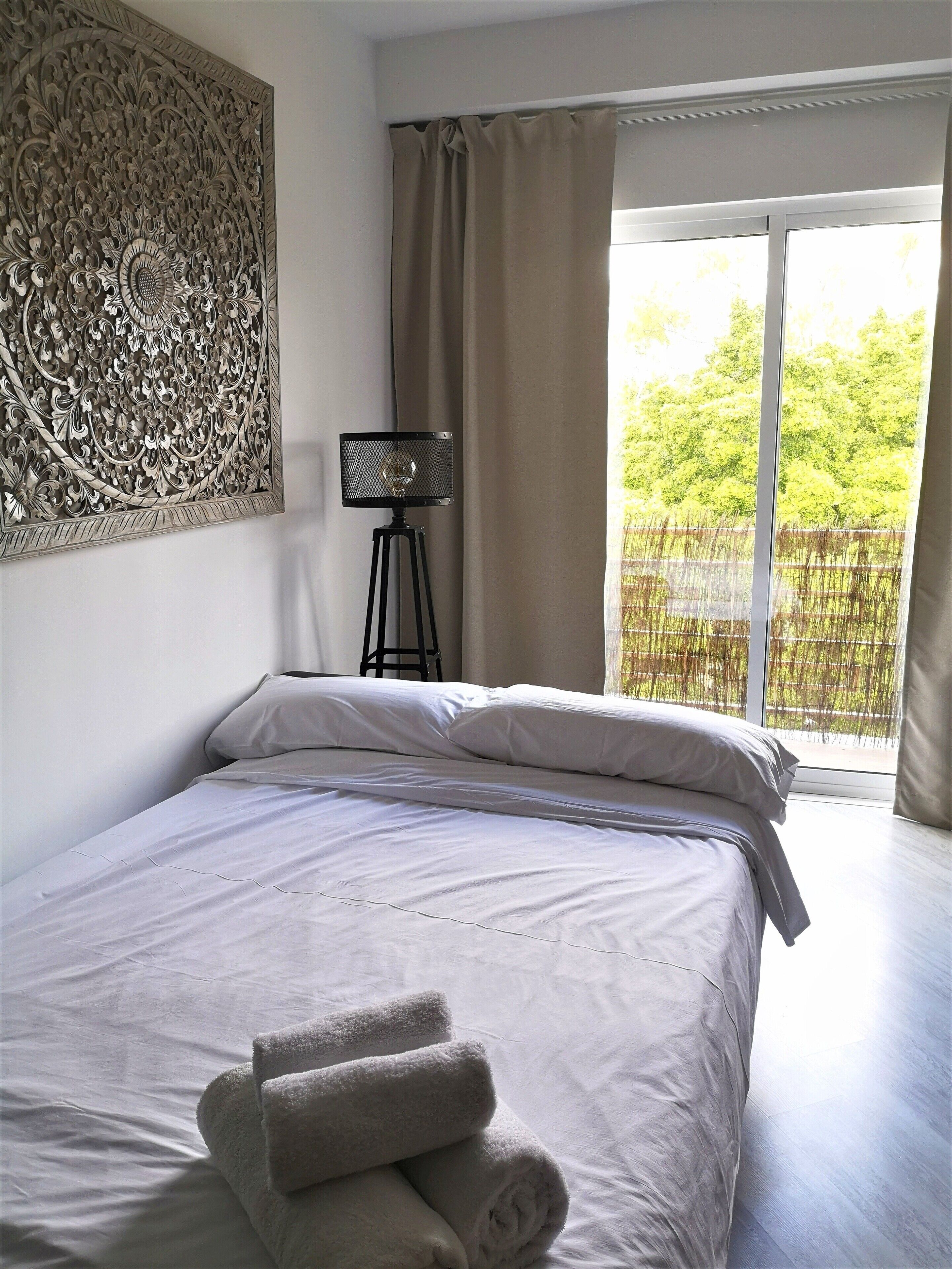 Un lit bien fait, avec du linge de lit blanc et des serviettes pliées, un élément décoratif mural et une vue sur la verdure par une grande fenêtre.
