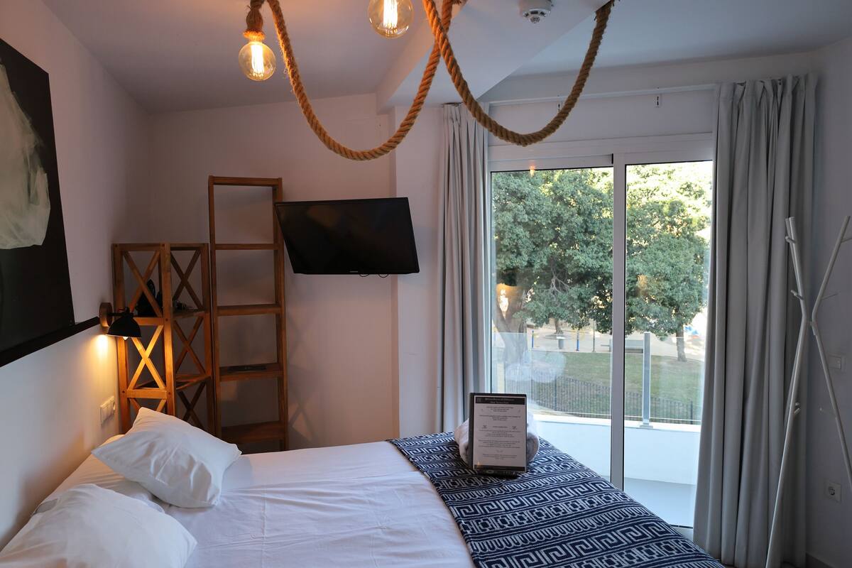 A hotel room with a bed, a mounted TV, a window with curtains, and a view of the outdoors.