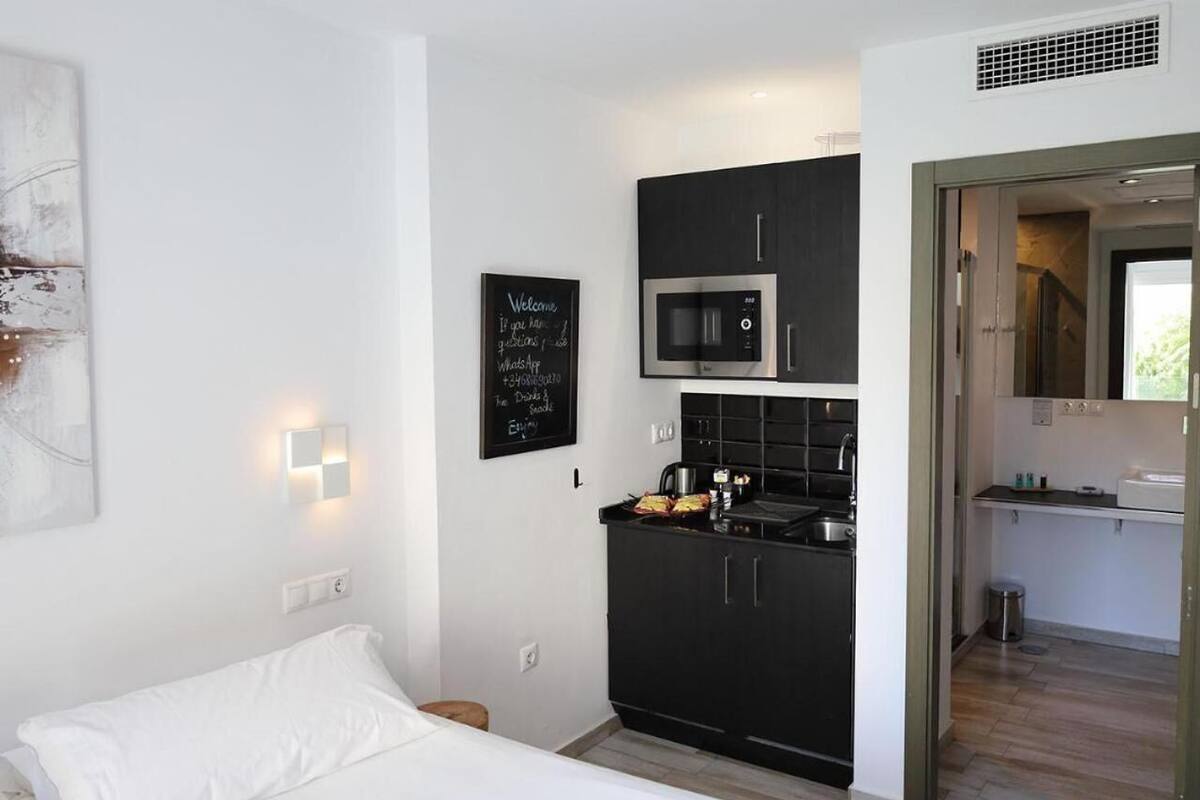 A compact hotel room with a kitchenette, a bed, a wall-mounted light, and a door leading to a bathroom.