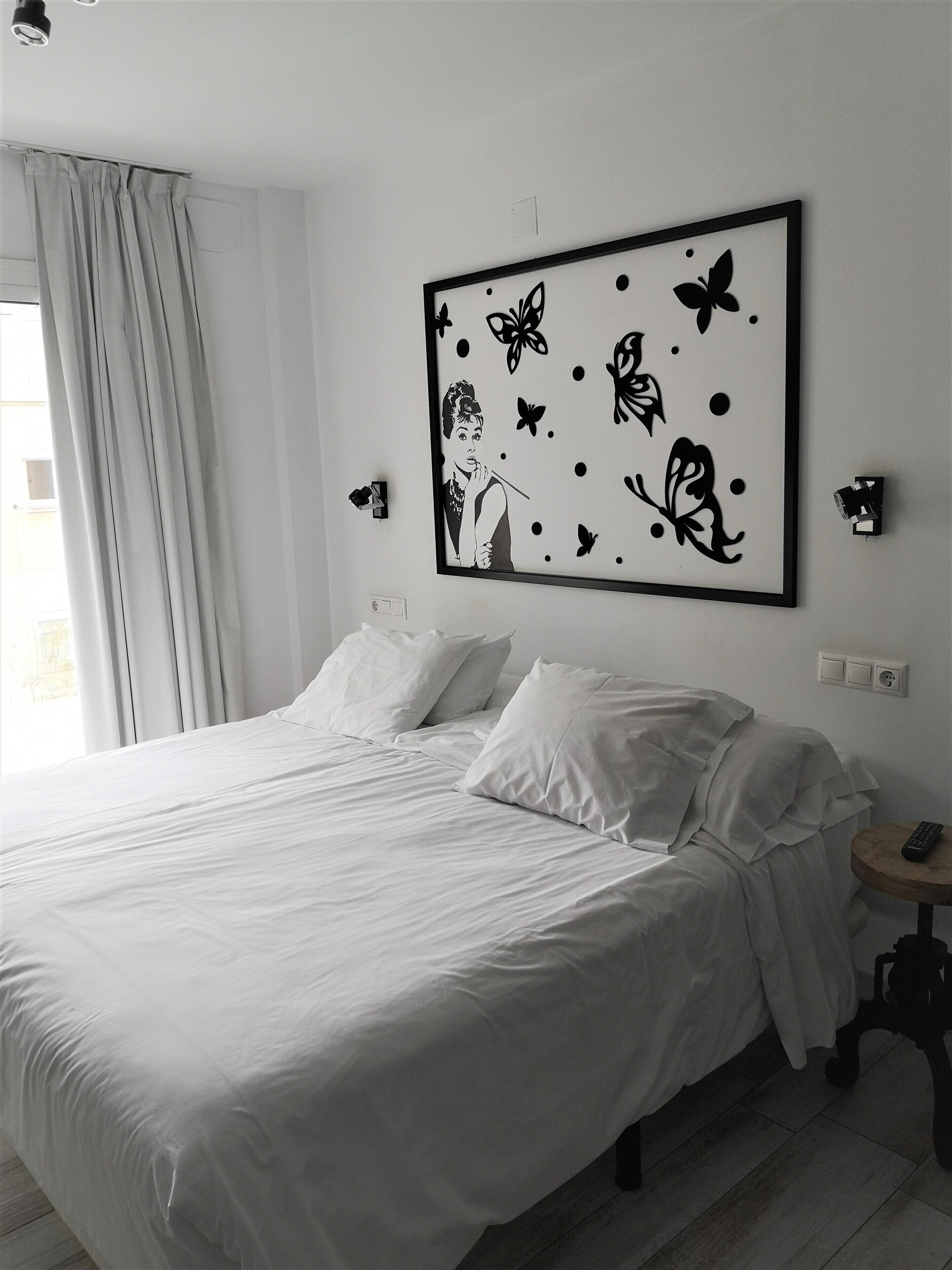 Hotelkamer met een groot bed, witte beddengoed, een kunstwerk aan de muur van een vrouw met vlinders, een nachtkastje met een afstandsbediening en een raam met gordijnen.
