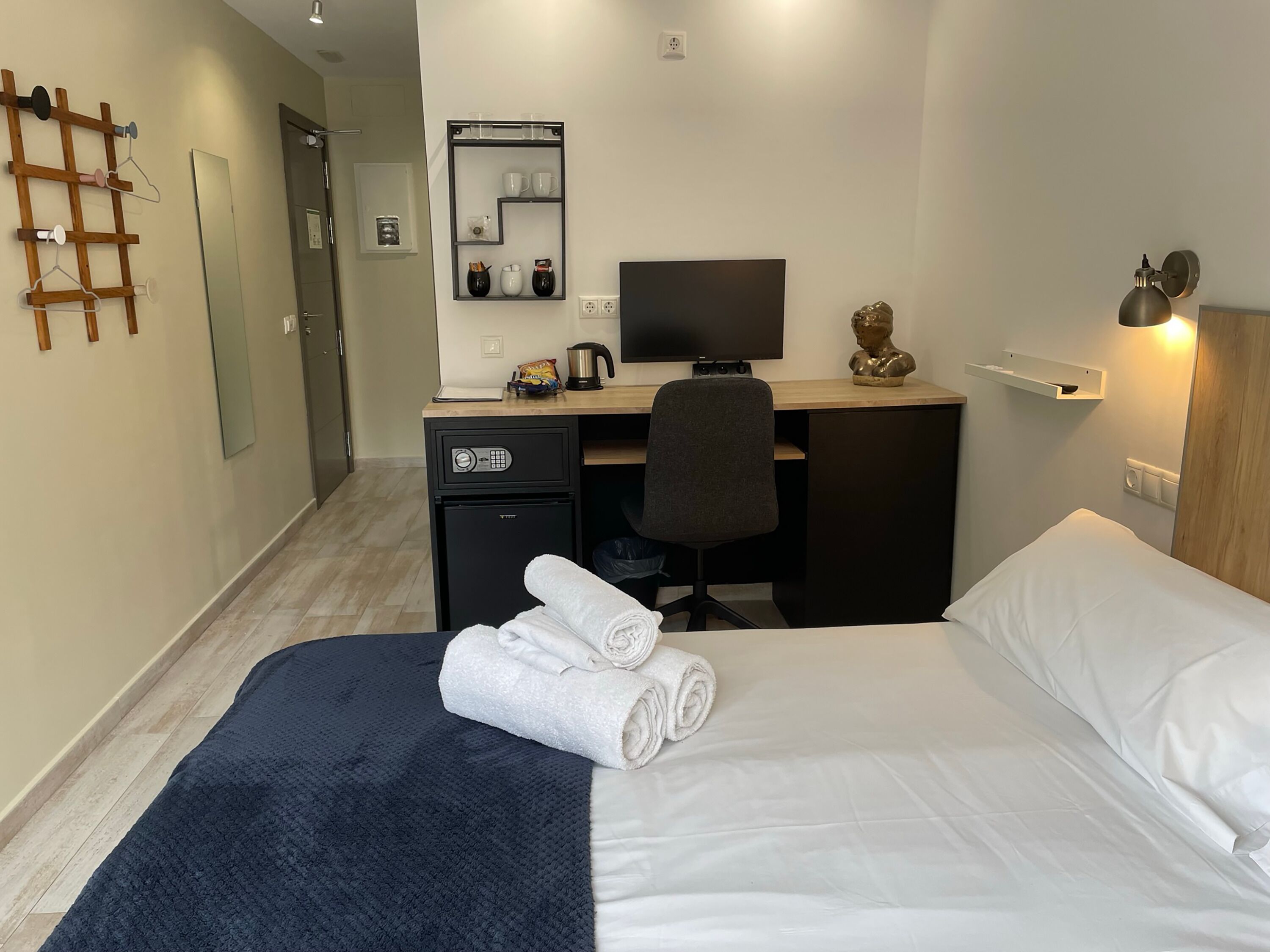 Habitación de hotel con una cama, un escritorio con computadora, una silla, una caja de seguridad pequeña y un estante con tazas y un hervidor.