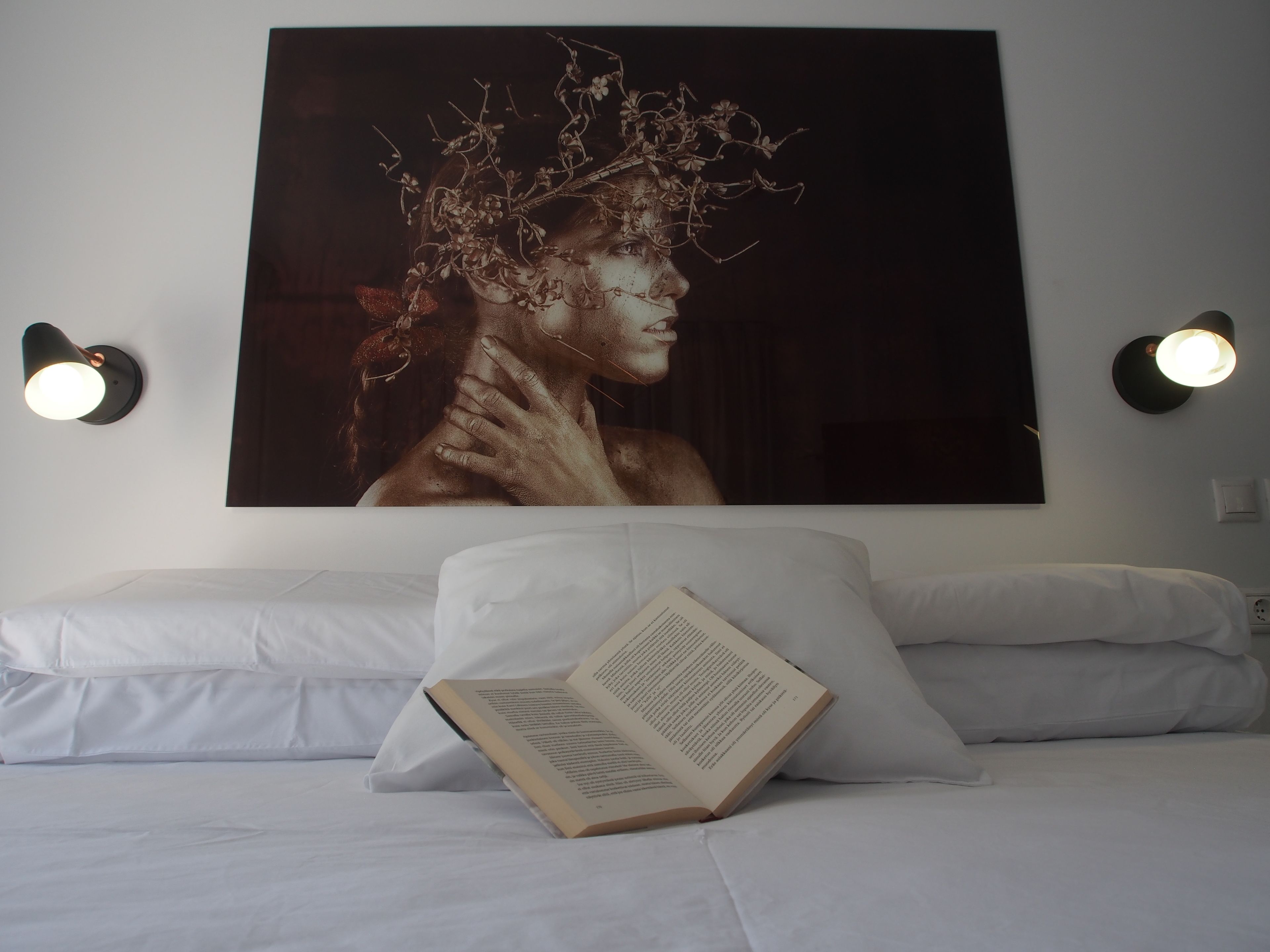 A bedroom with a large framed artwork on the wall, a bedside table with a book, and two wall-mounted lamps.