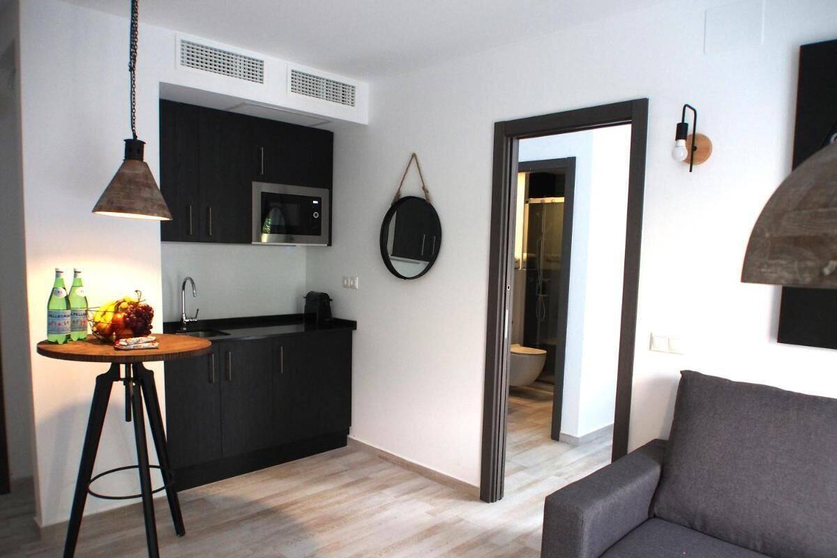 A modern living room with a grey sofa, a kitchen area with dark cabinets and a microwave, and a dining table with bottles and fruit.