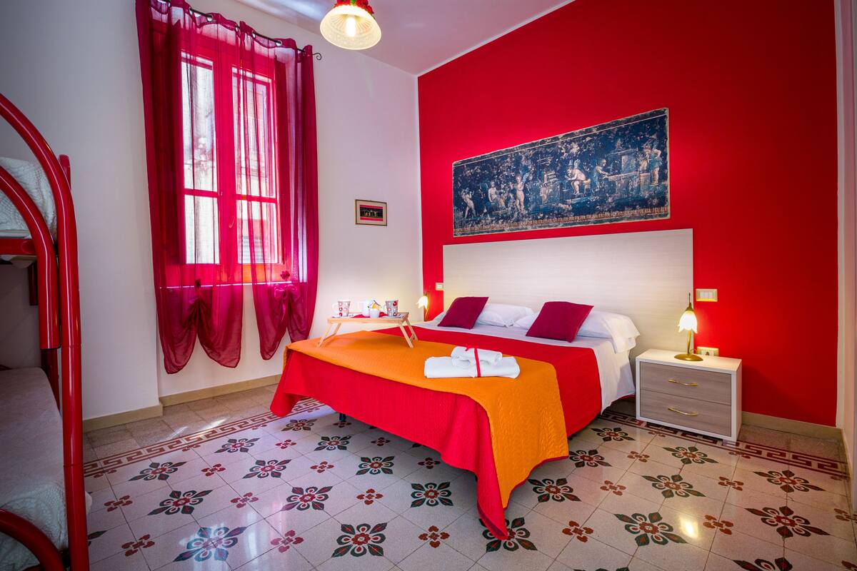 A room with a bed, a nightstand, a table with a red tablecloth, a window with red curtains, and a red wall with a painting.