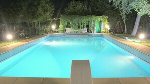 Una piscina rectangular con superficie de agua azul cristalina, rodeada por un suelo pavimentado. Hay dos sillas de descanso y una mesita bajo una pérgola con follaje verde. El área está iluminada por varias luces en el suelo.