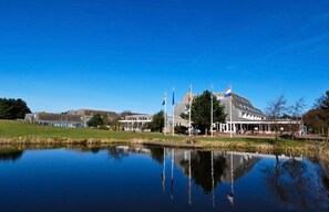 A lakeside building with a reflection in the water, surrounded by greenery and flags.