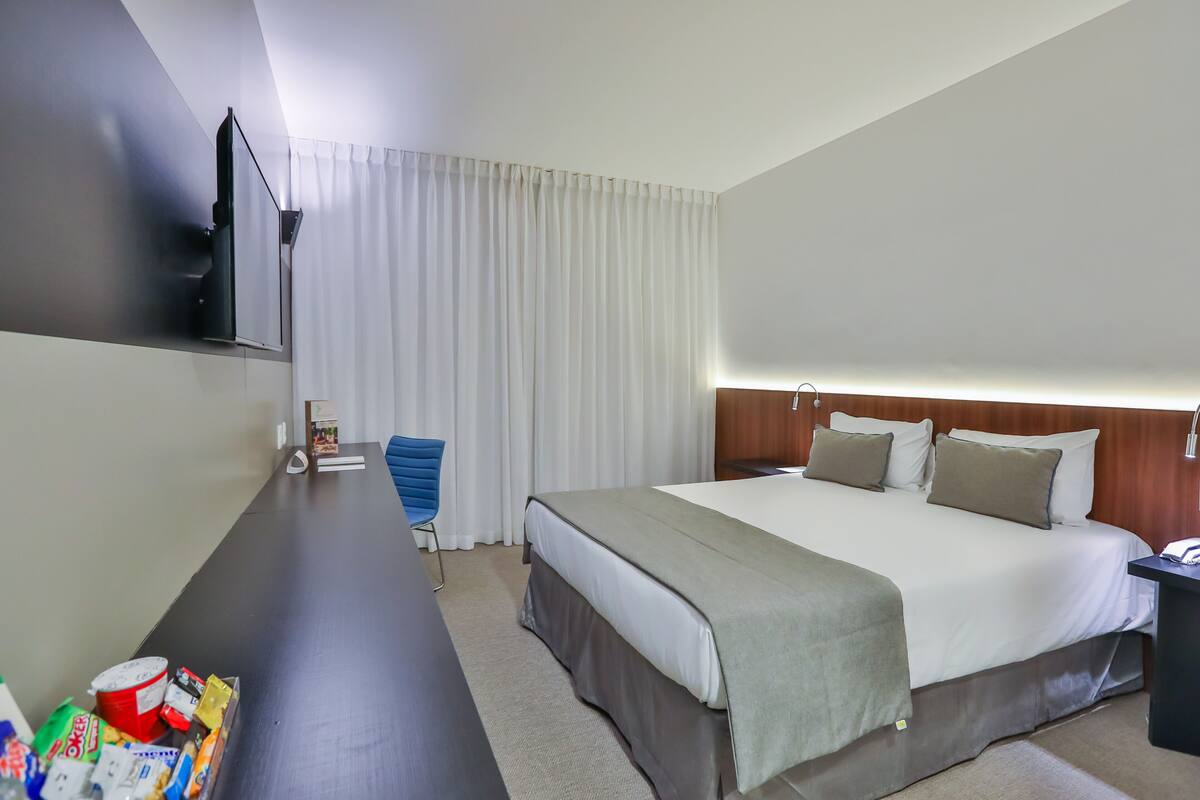Habitación de hotel con una cama grande, un escritorio con una silla, un televisor montado en la pared y una ventana con cortinas.