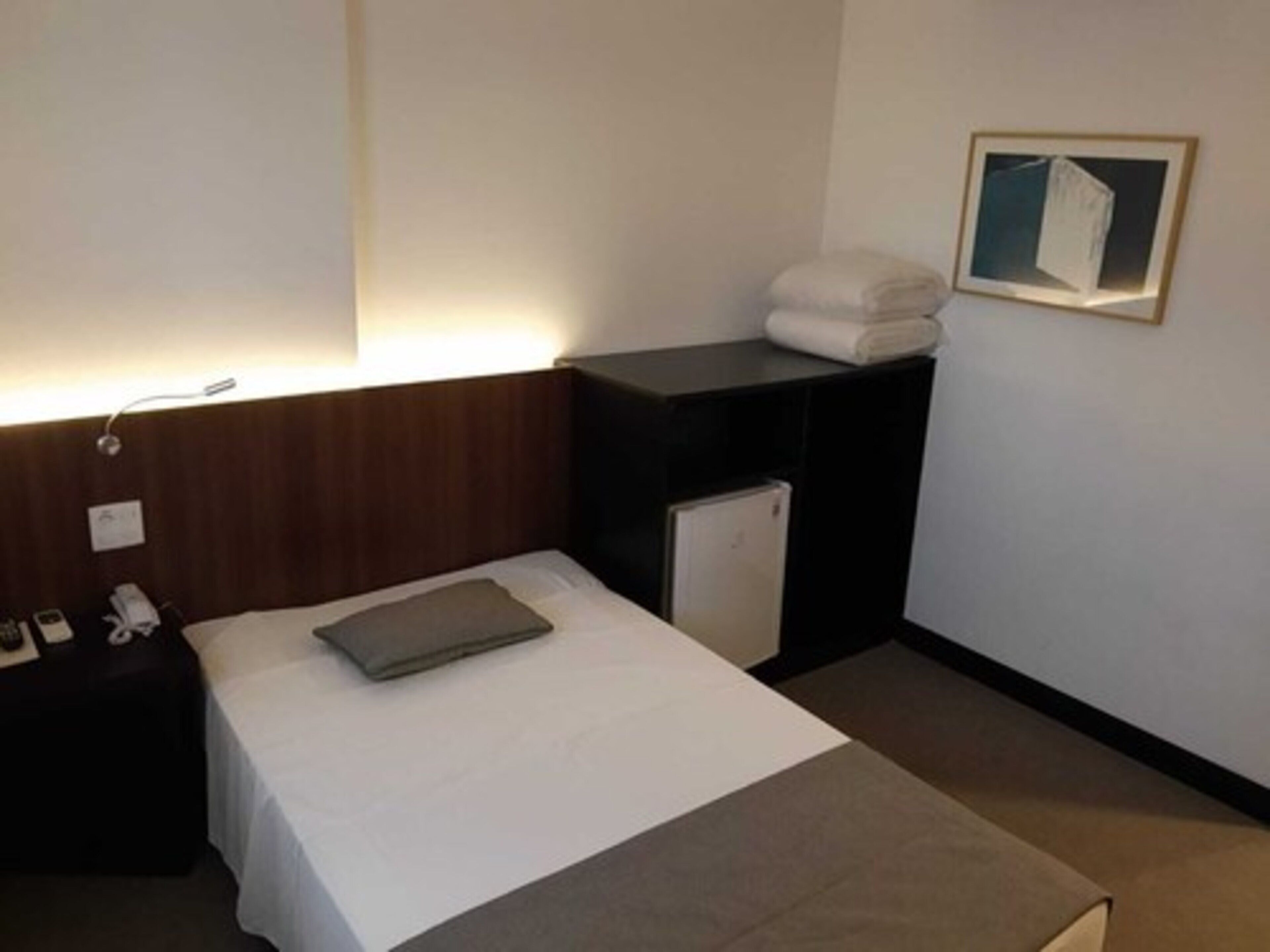 A hotel room with a bed, a nightstand, a small refrigerator, and a wall-mounted shelf with towels.